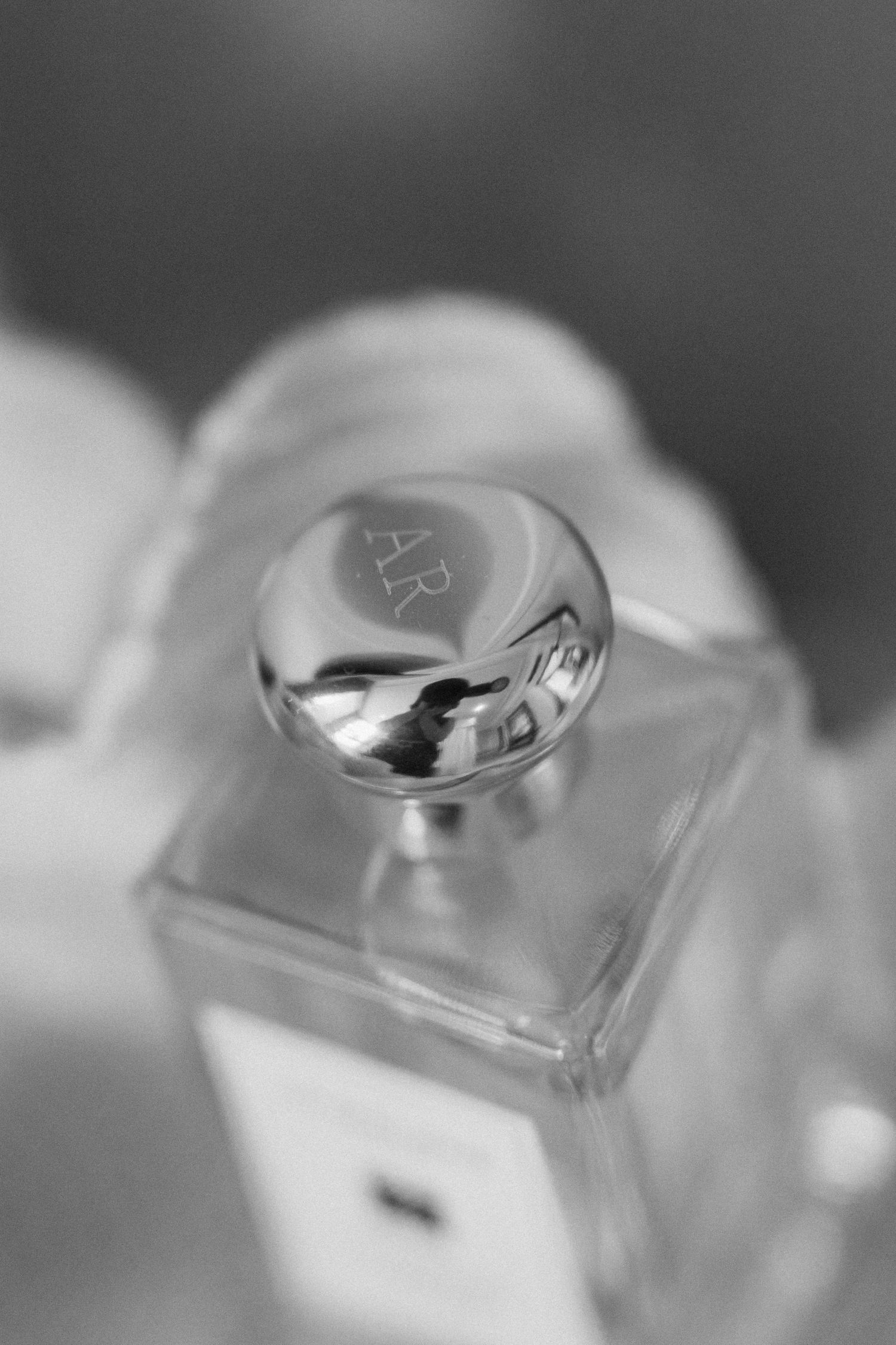 Close-up black and white macro shot of engraved silver perfume bottle cap with initials on square glass bottle.
