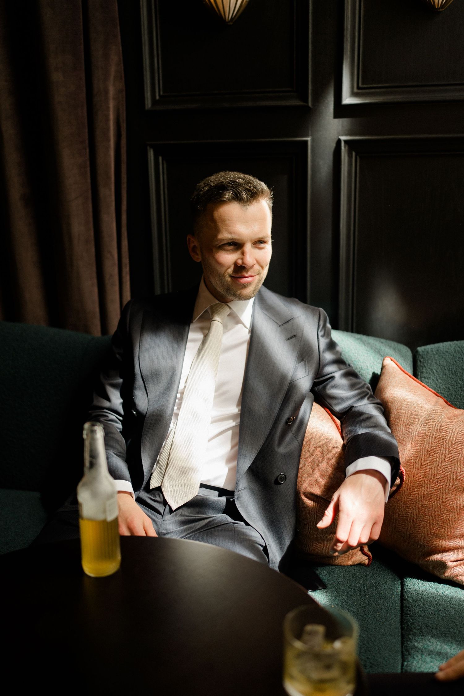 Elegant man in gray suit relaxing on a green velvet sofa with a beer bottle on a dark table.