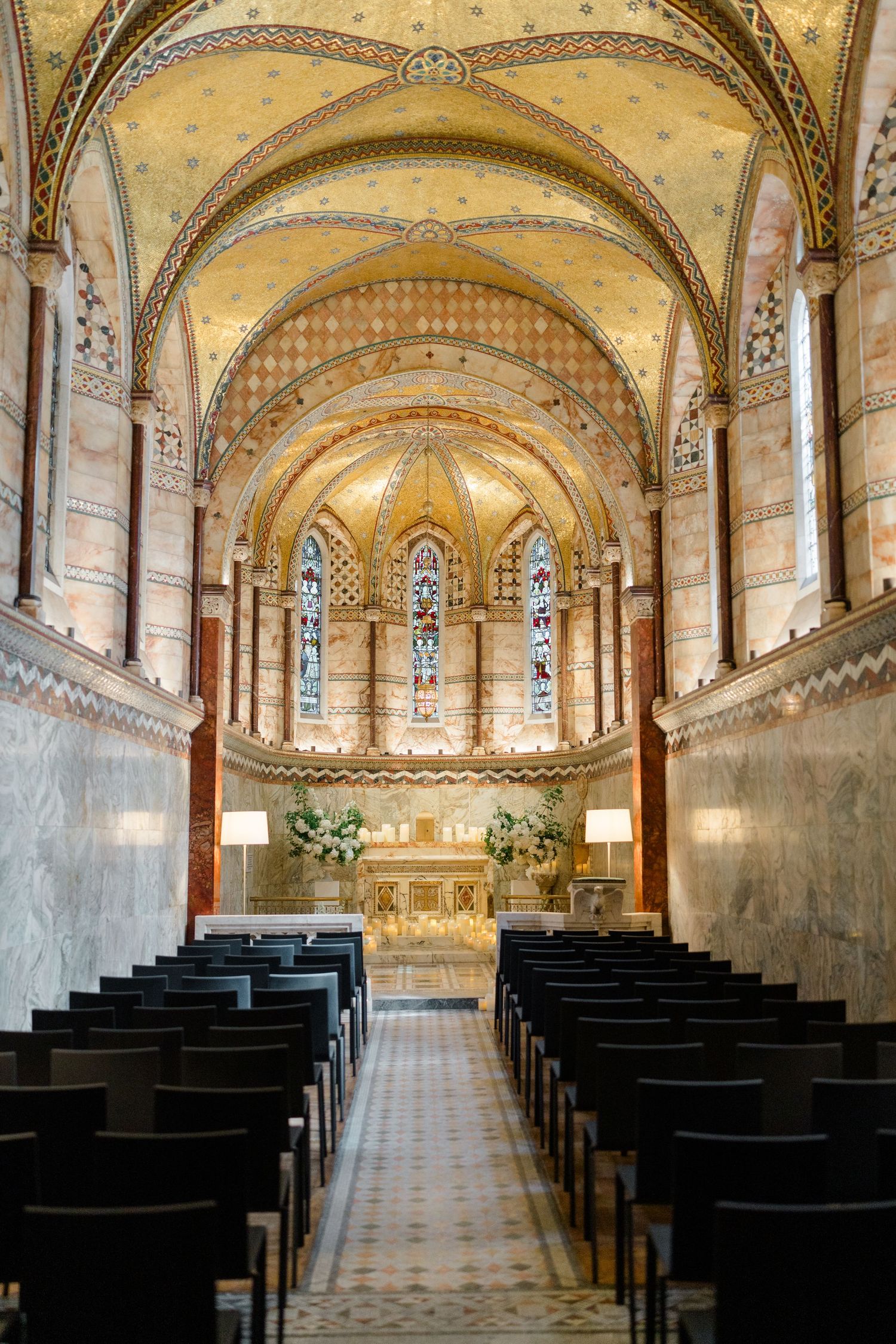 Elegant chapel interior with golden vaulted ceiling, stained glass windows, and white floral altar arrangements for a wedding ceremony.