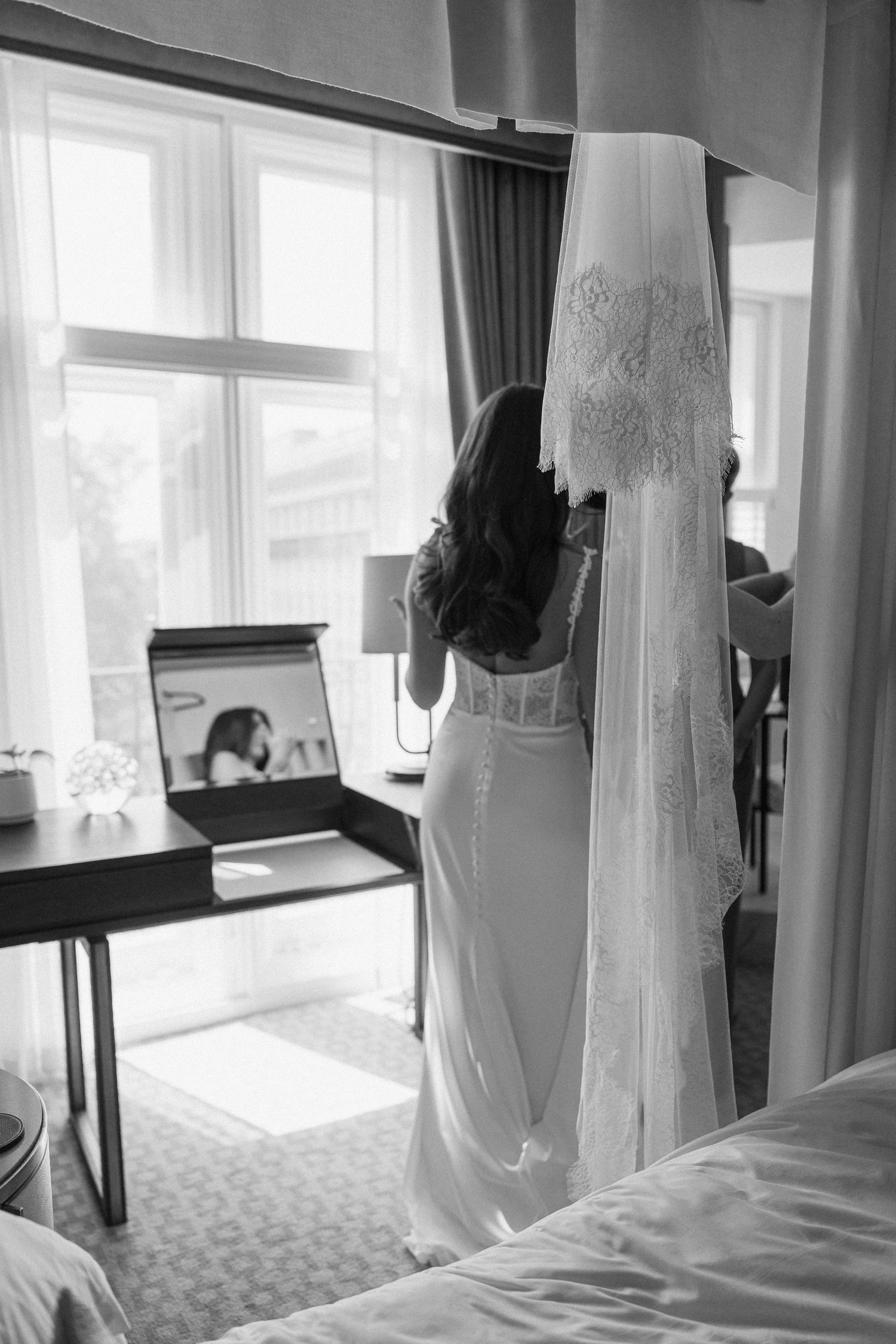 Black and white photo of a bride in a wedding dress standing by a window with her veil hanging nearby.