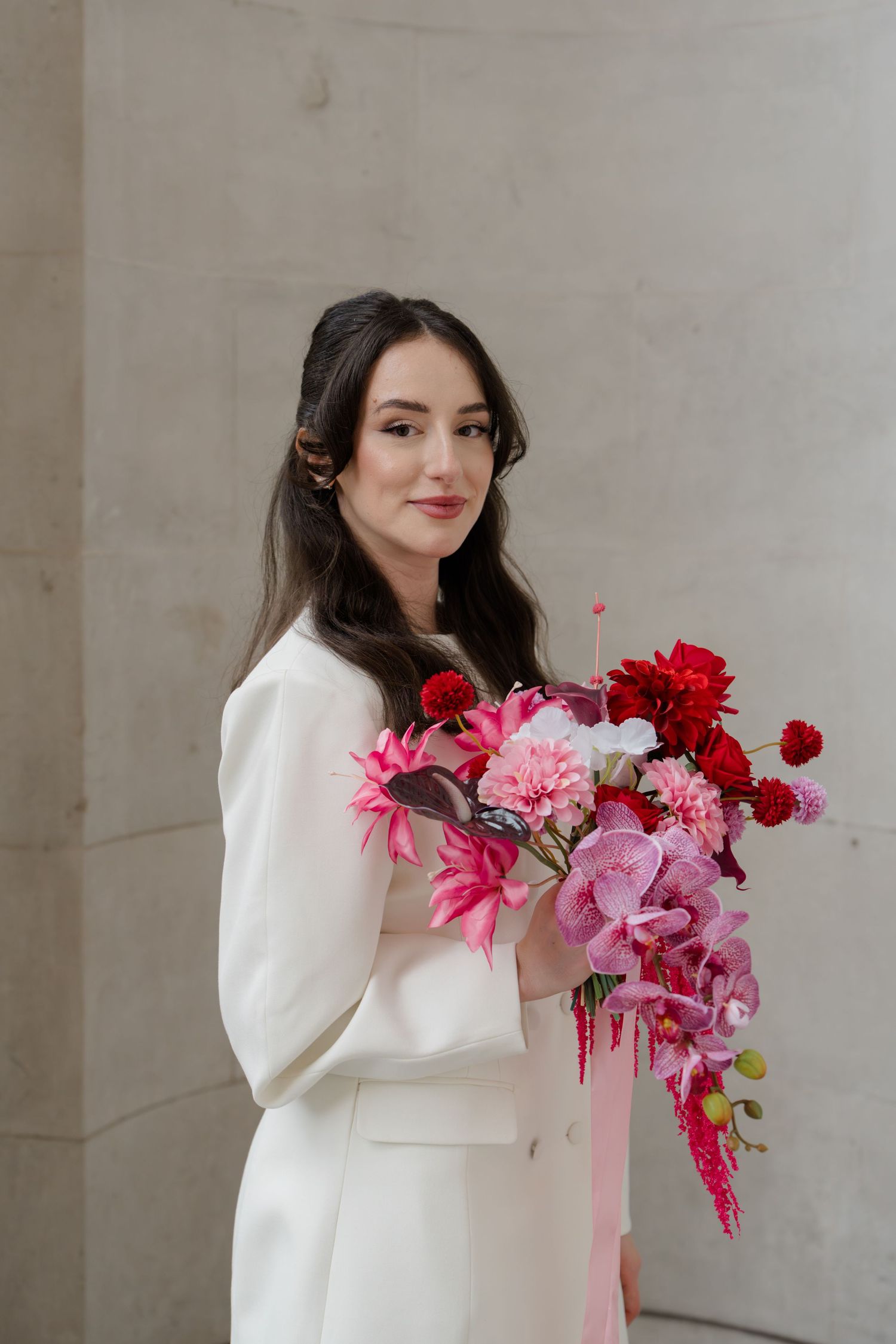 Elegant bride in white suit holding vibrant pink and red floral bouquet with orchids and carnations.
