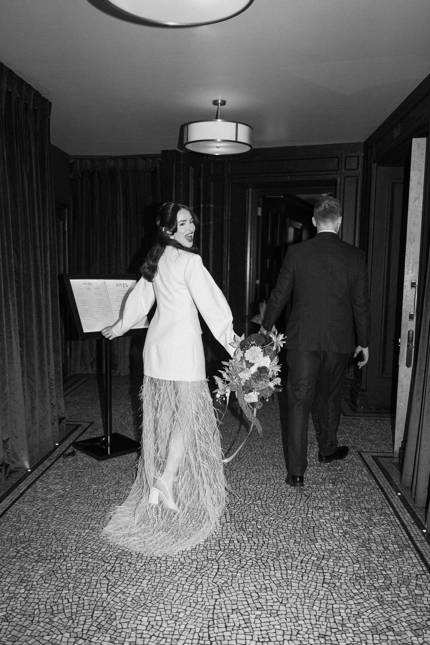 Black and white photo of bride in white blazer and feather skirt holding bouquet walking with groom through hotel corridor.
