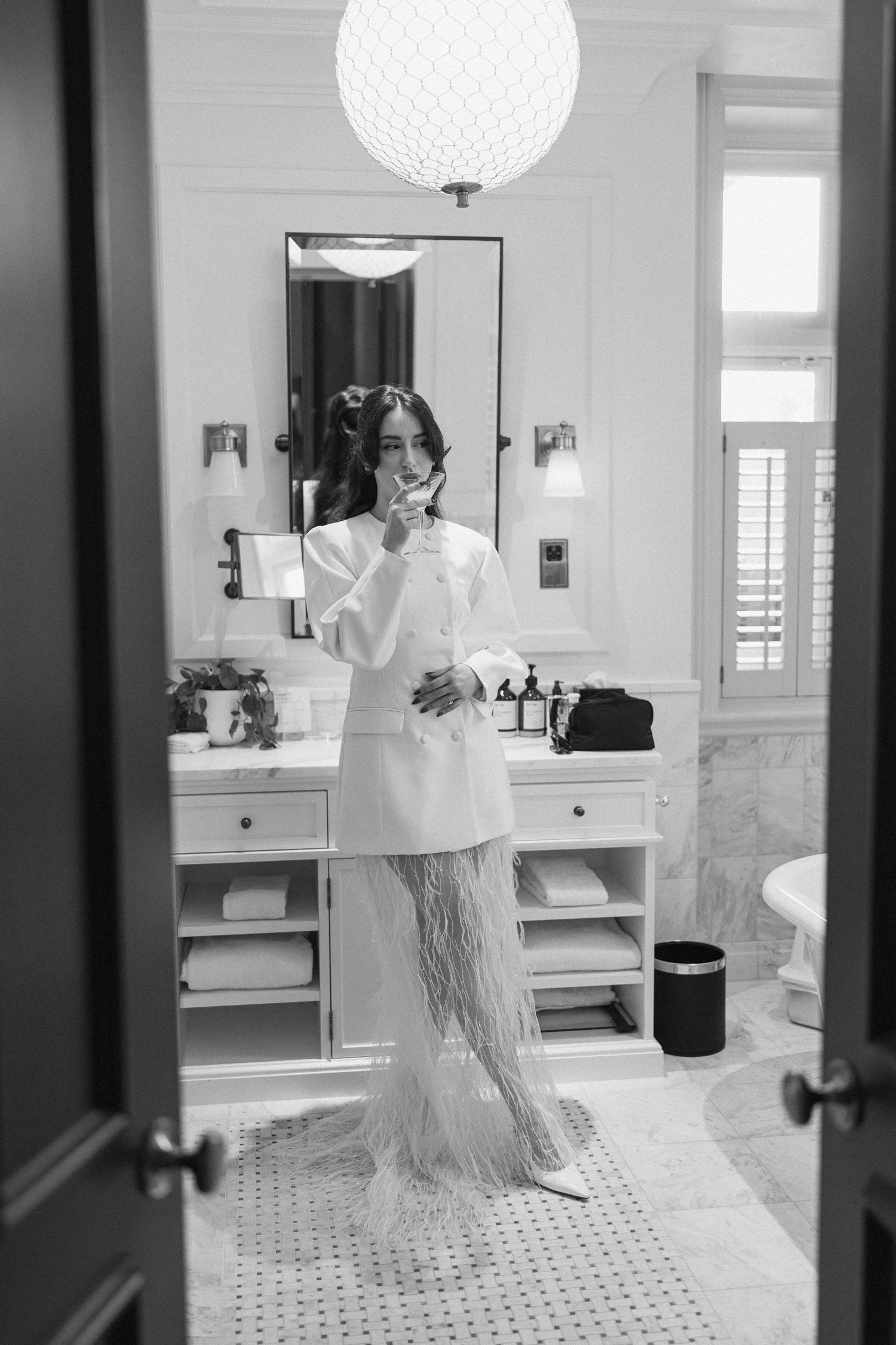 Bride in white feather wedding outfit applying makeup in elegant marble hotel bathroom mirror.