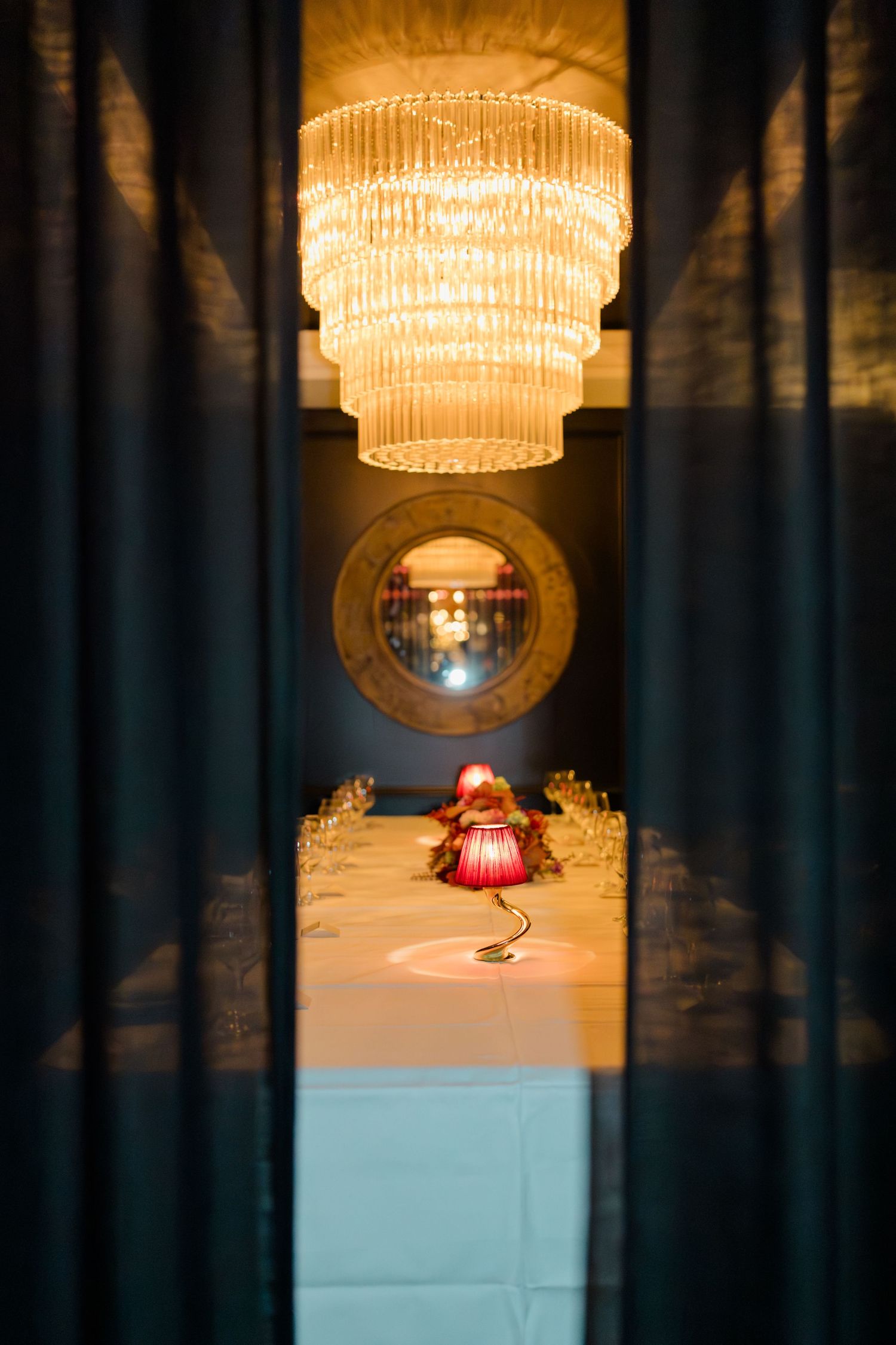 Elegant dining room viewed through curtains with tiered crystal chandelier and red lamp centerpiece.