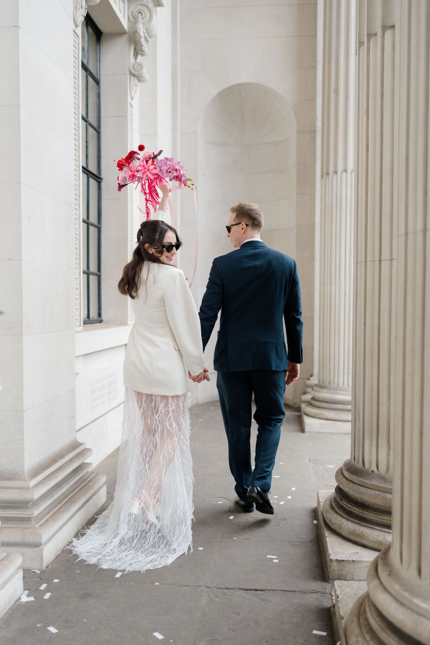 Bride in white blazer and lace skirt walks with groom past stone columns.