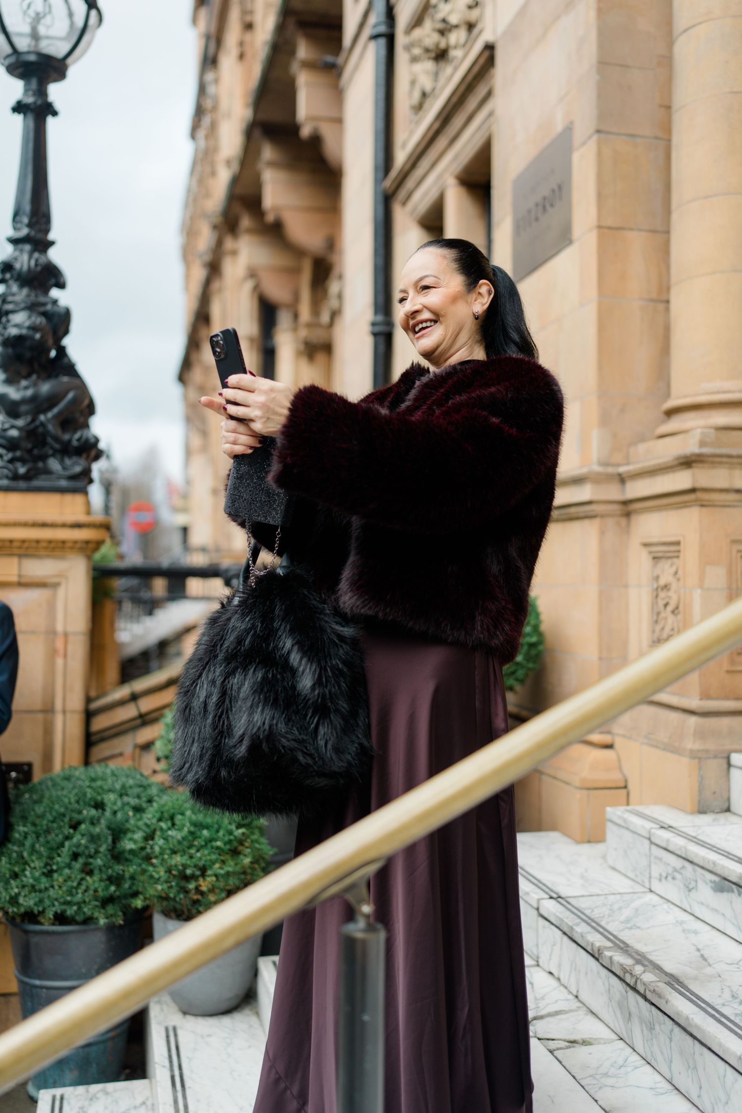 Smiling woman in burgundy fur coat takes selfie on grand building steps holding black fur bag.