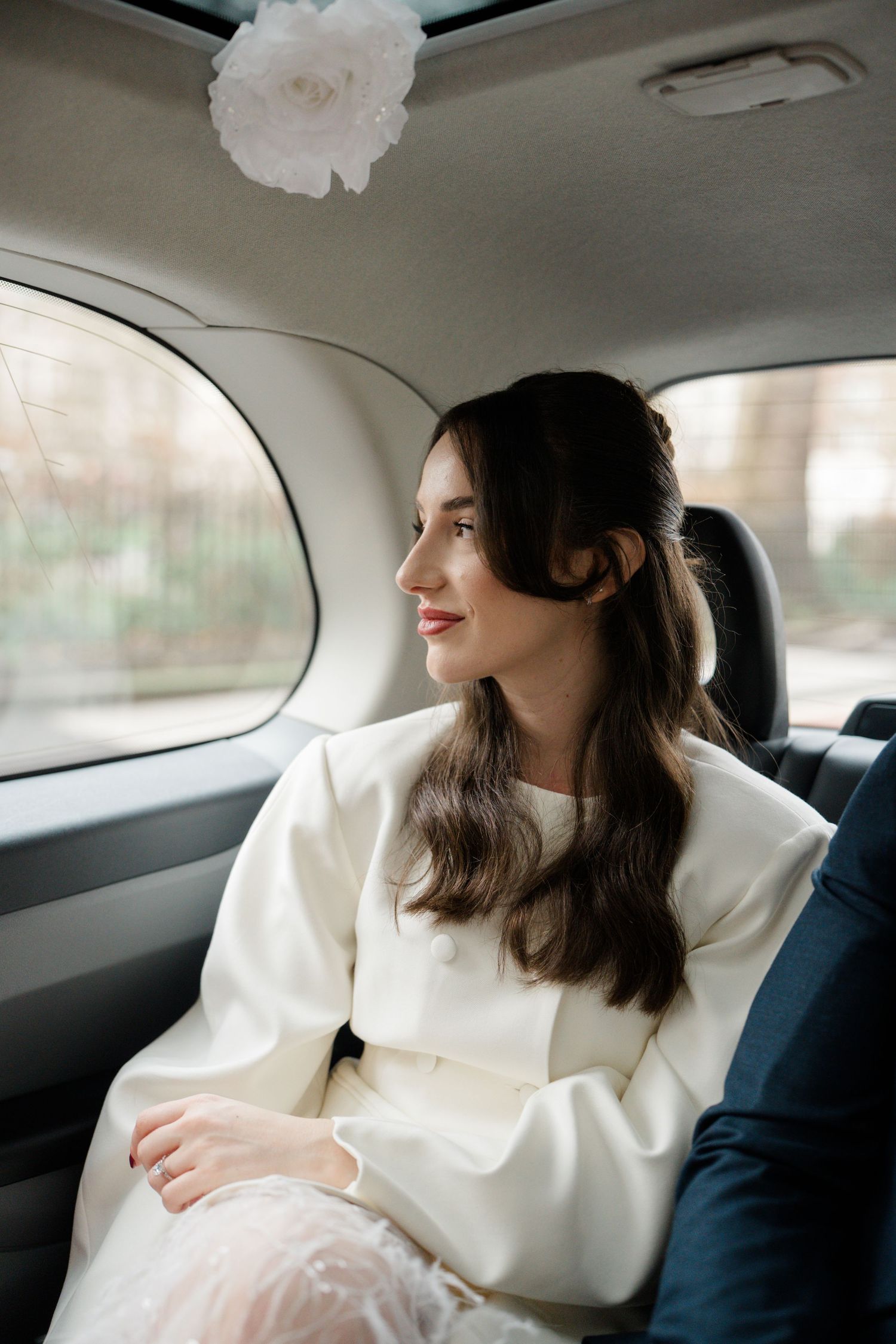 Bride in white dress sitting in car backseat, smiling out window with floral decoration above.