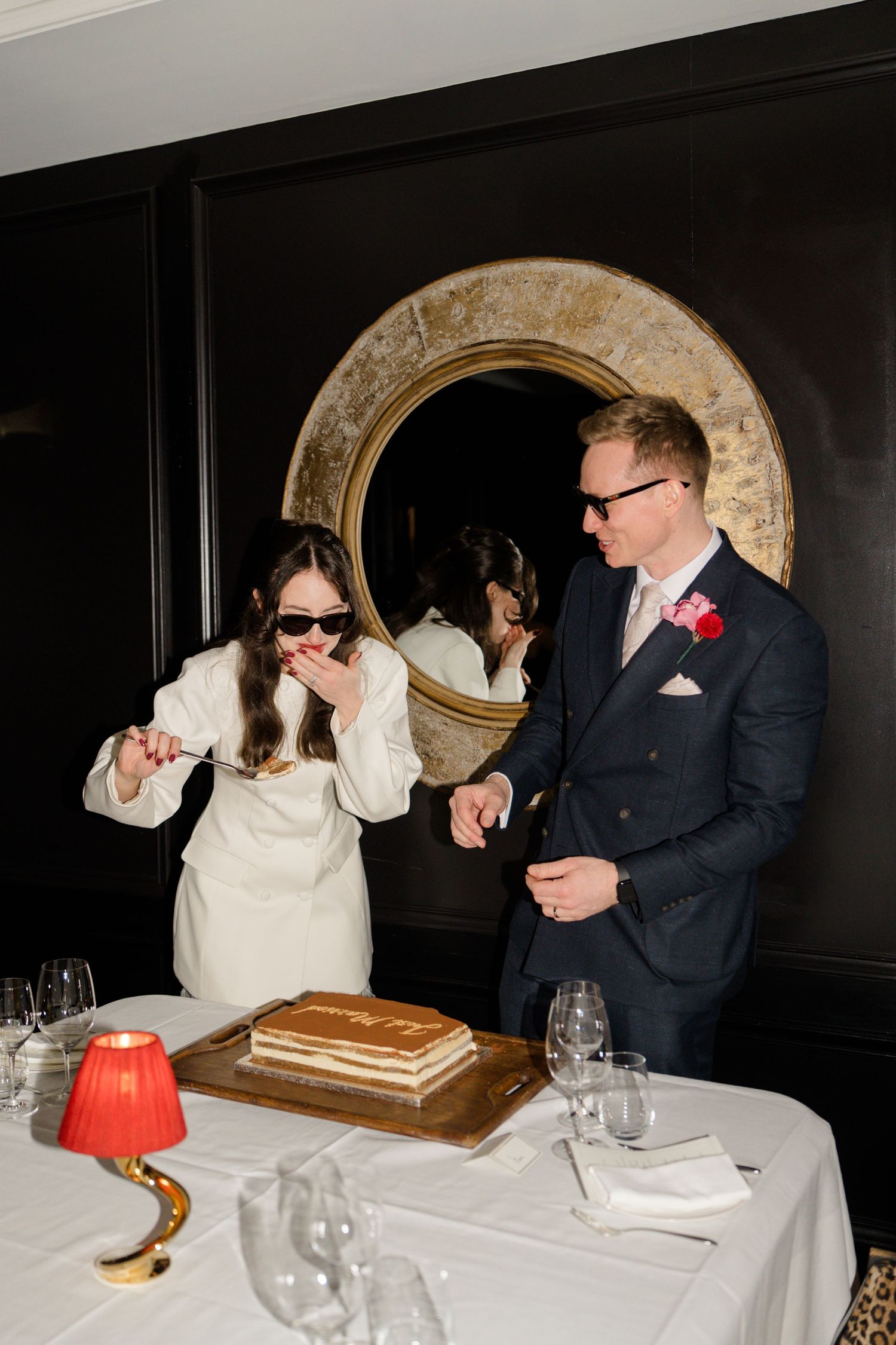 Bride in white dress and sunglasses tastes wedding cake beside groom in navy suit with floral boutonniere.