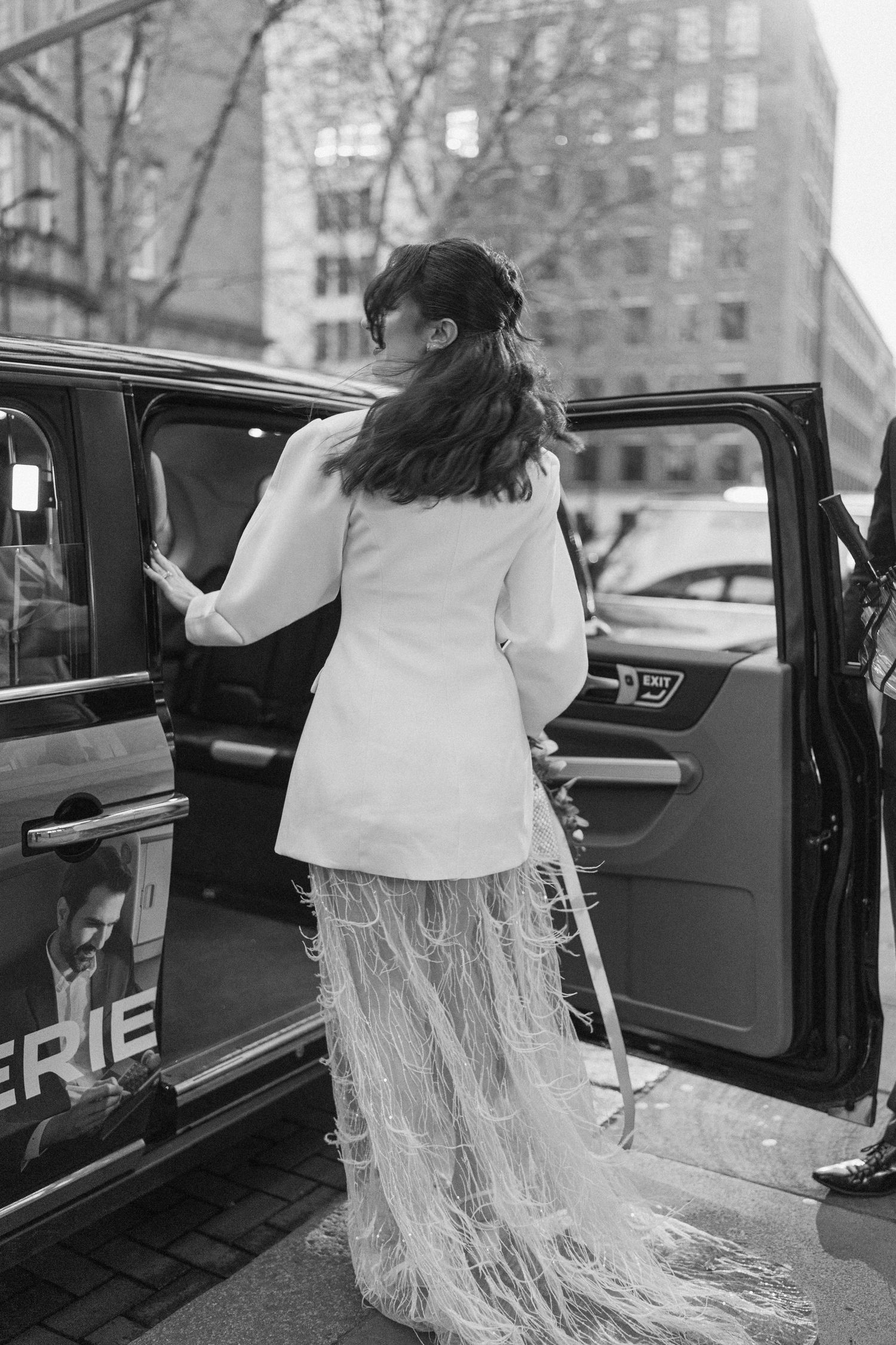 Black and white photo of bride in white feathered suit getting into black London taxi cab.