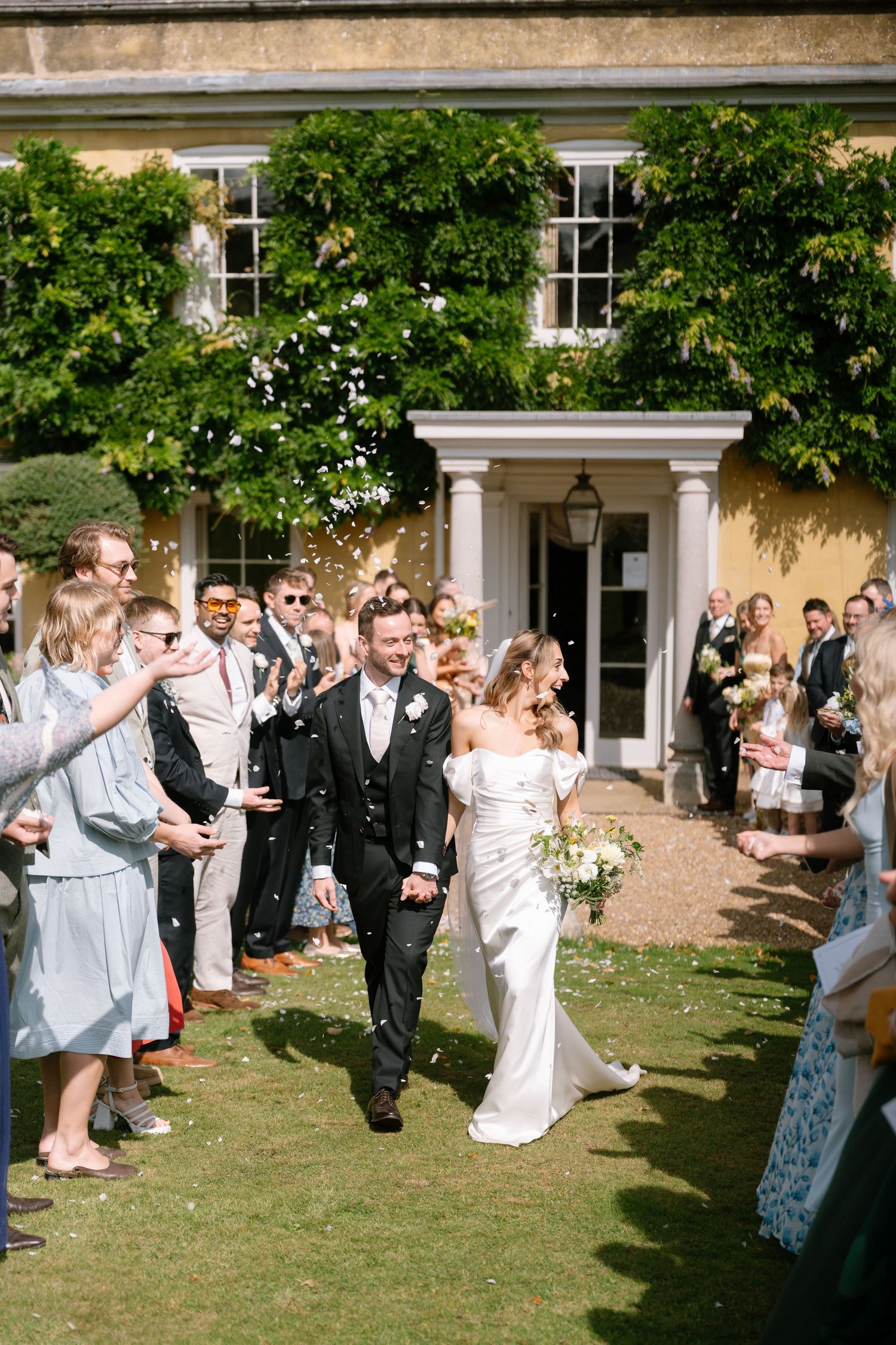 Wedding guests line up outside a stately venue to celebrate with confetti on a sunny day.