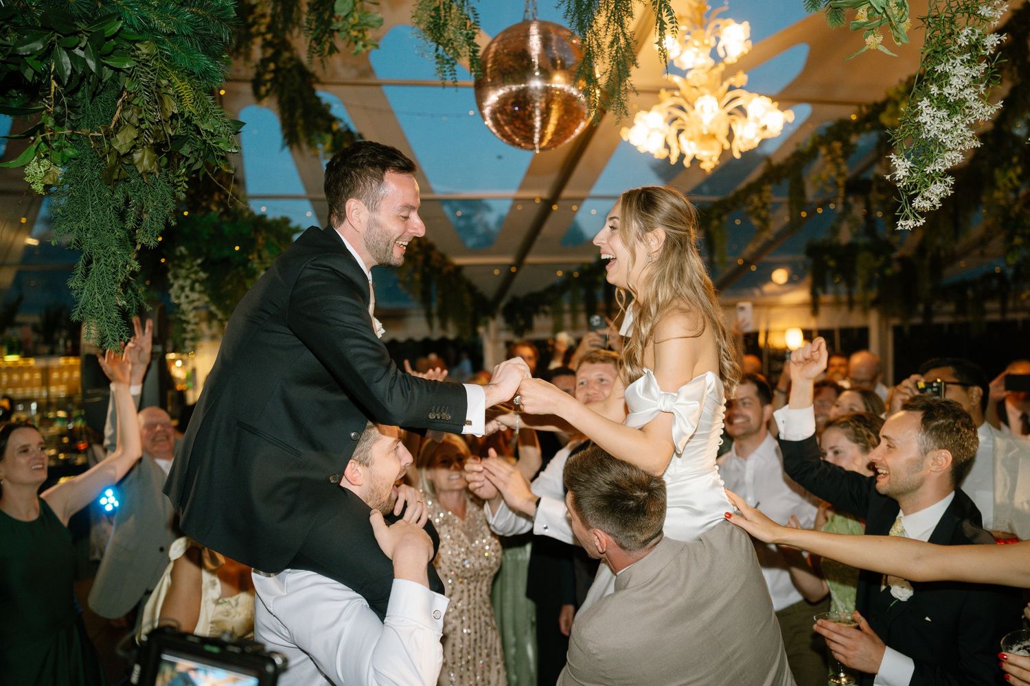 Festive wedding party dancing beneath hanging greenery and warm lighting.