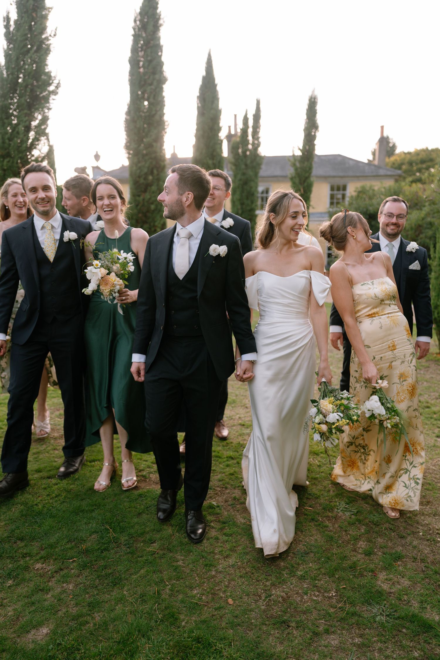 Wedding party walking and laughing together on grassy lawn with tall cypress trees in background.