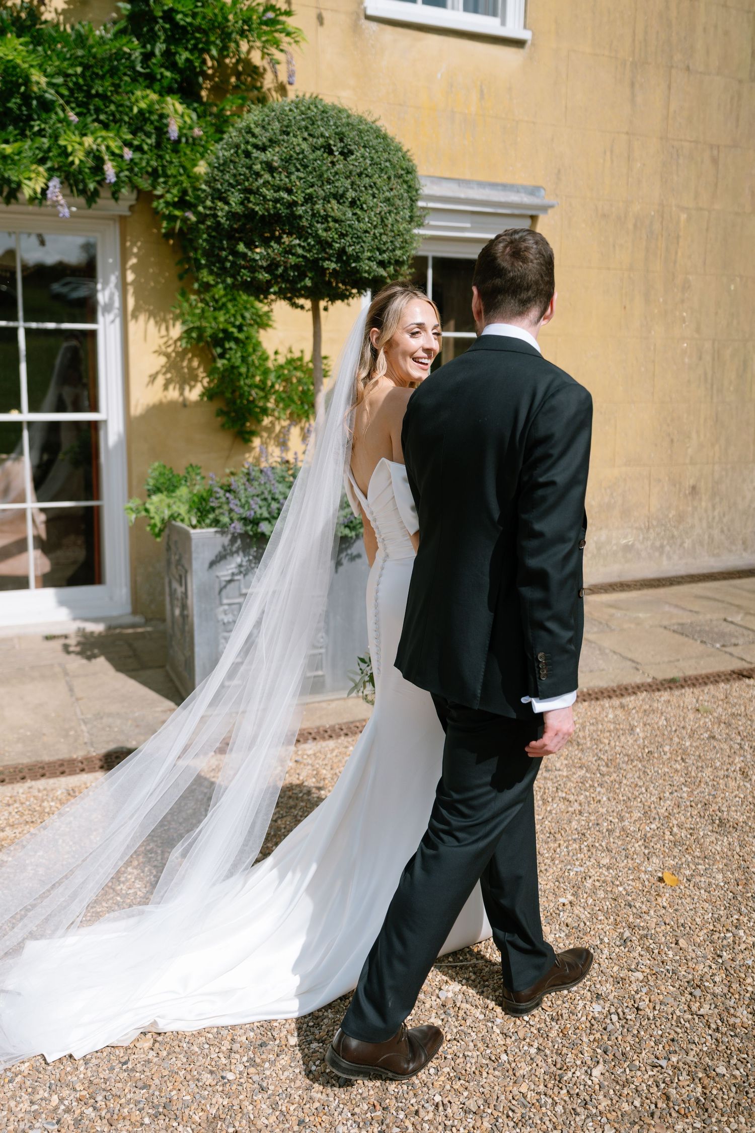 Wedding couple walking along a path beside a yellow stone building with trailing veil.