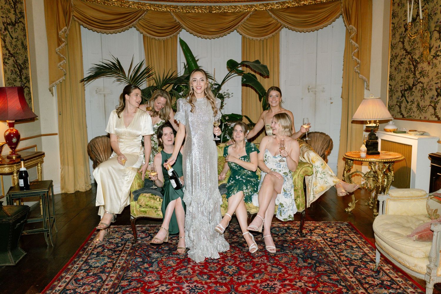 Wedding party in elegant dresses lounging on ornate furniture in a luxurious room with gold drapes.