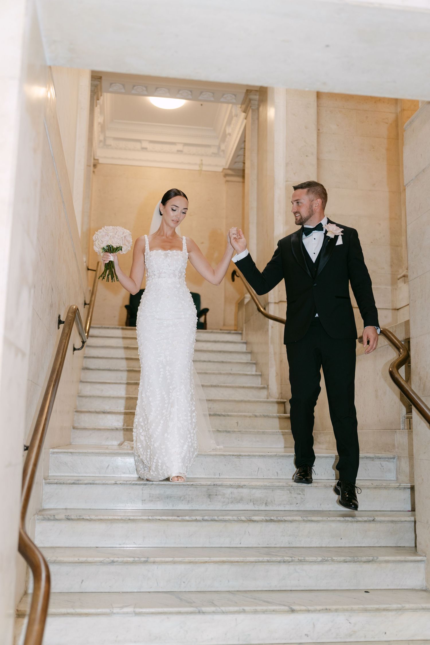 Happy couple walks down marble stairs holding hands in elegant wedding attire.