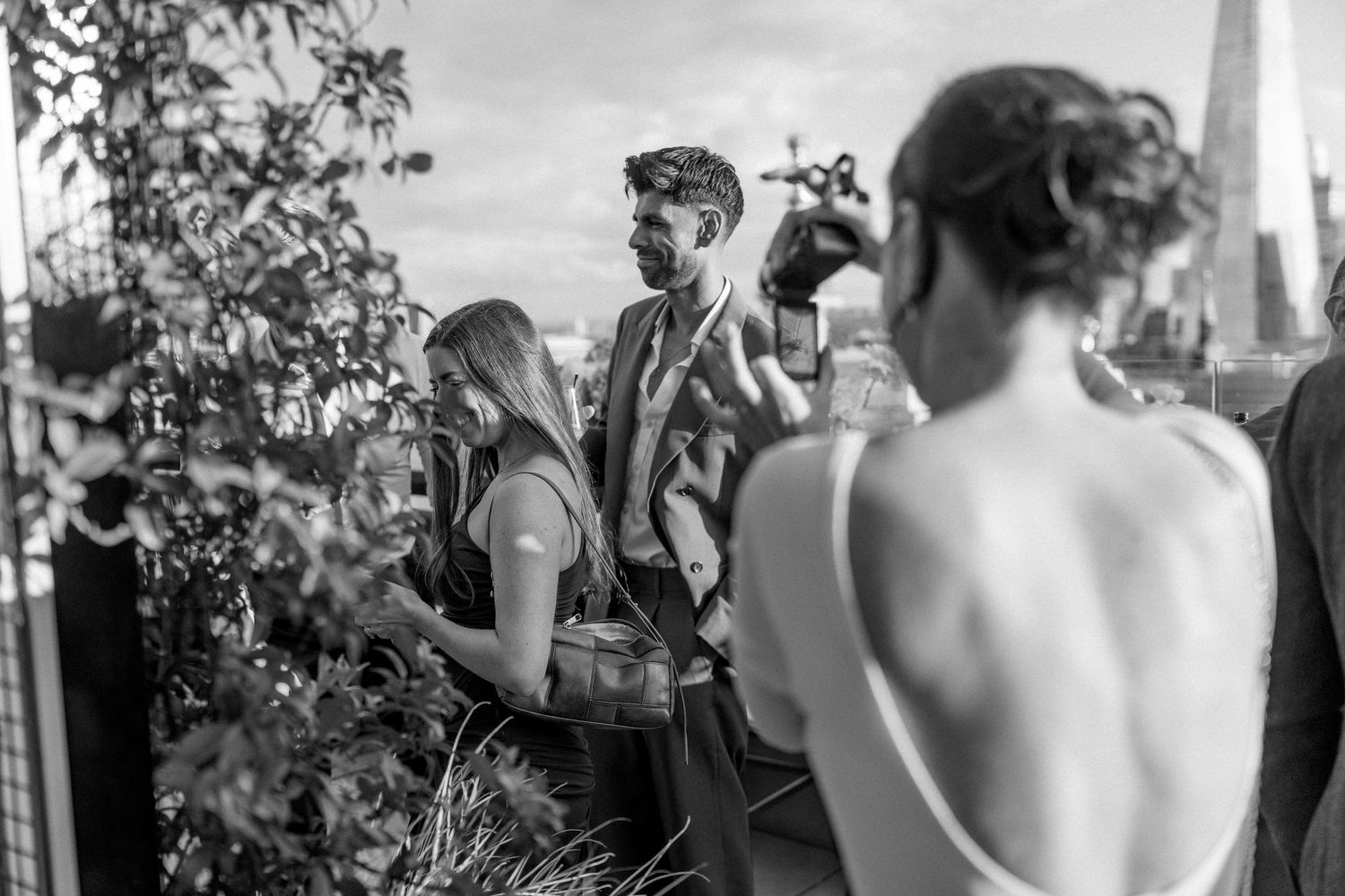 Black and white photo of wedding guests mingling on a rooftop with urban views.