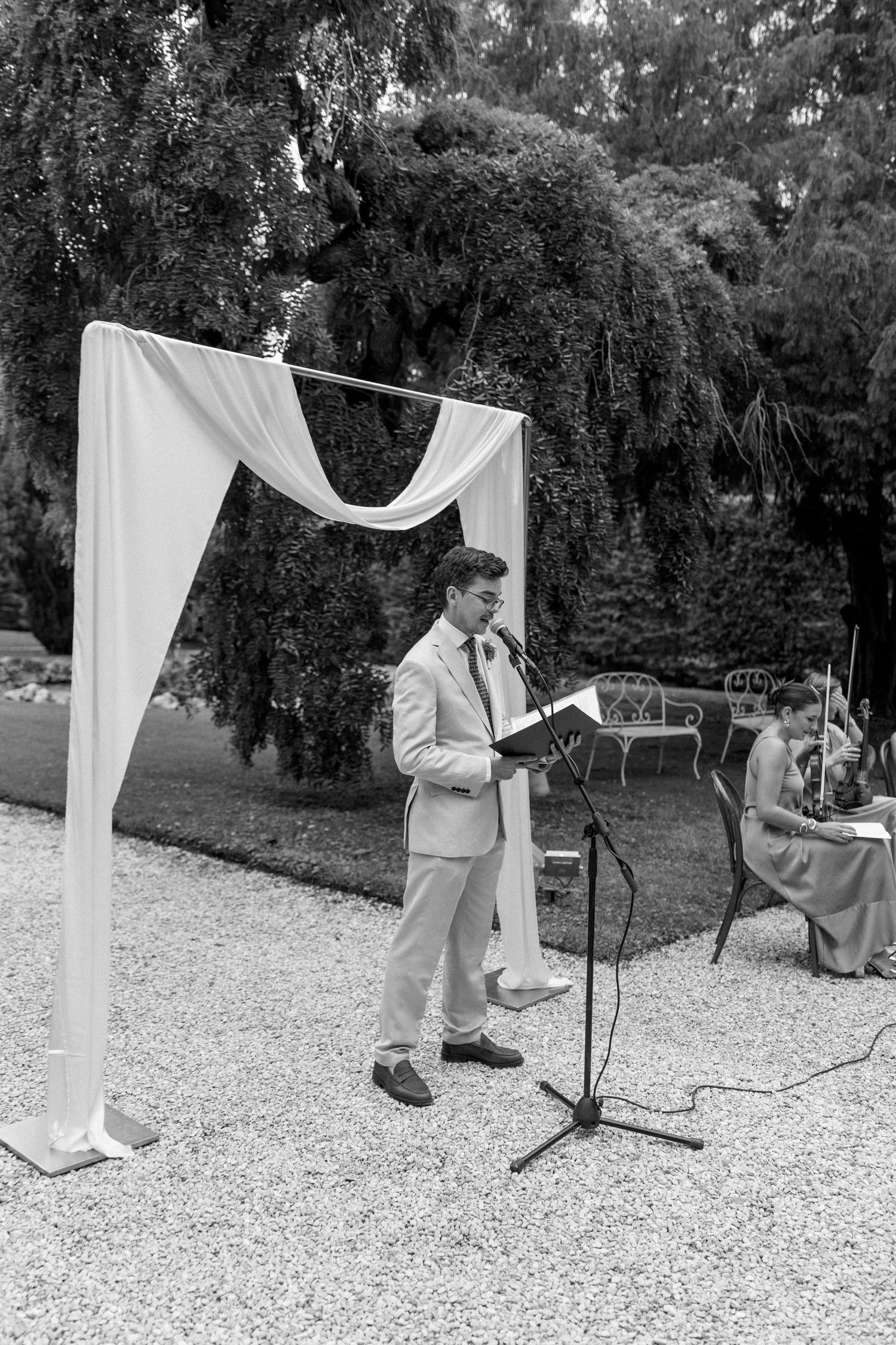 A wedding ceremony setup with white fabric draping and microphone stands on a gravel path.