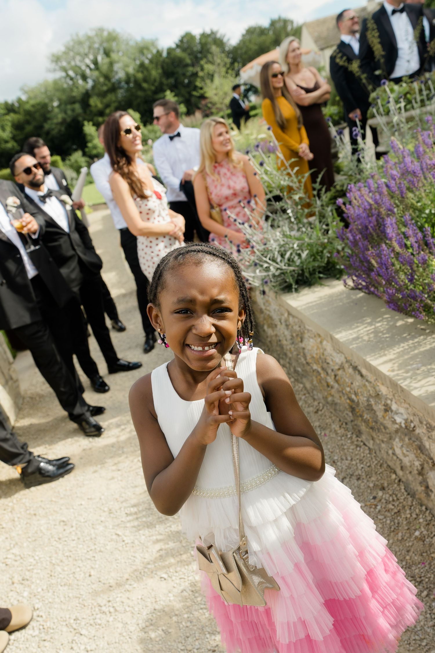 A joyful flower girl in a white and pink dress clasps her hands at an outdoor wedding ceremony.