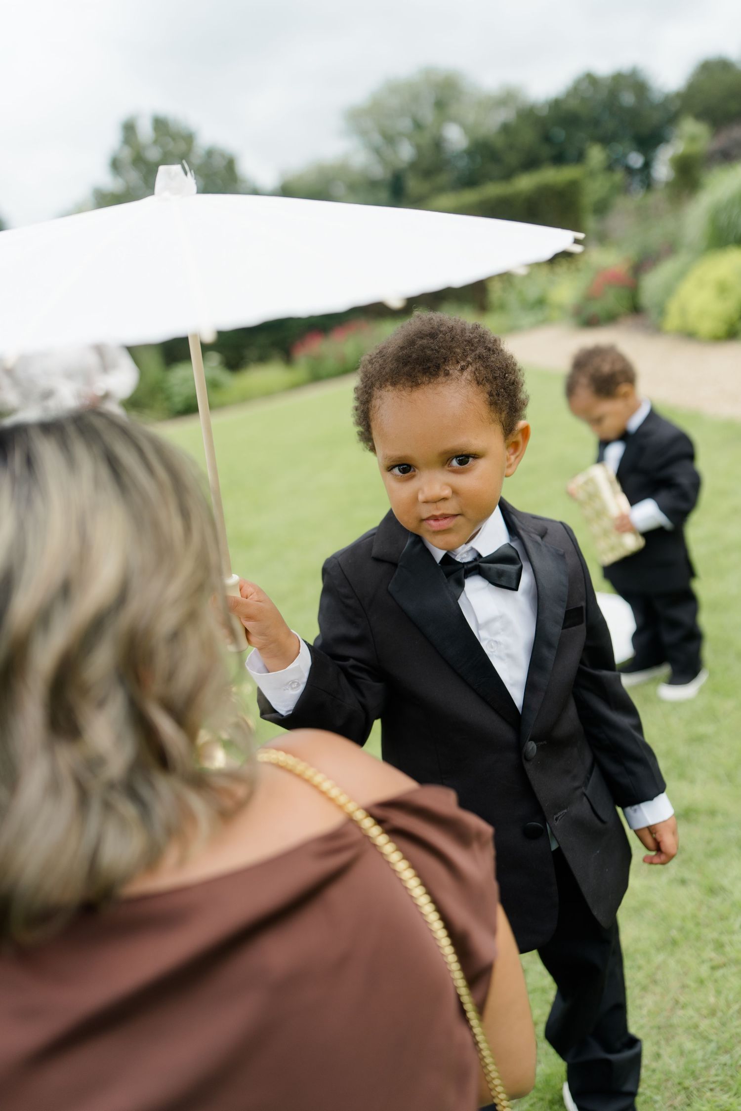 A young boy in a tuxedo holds a white parasol while standing on a garden lawn at a wedding.