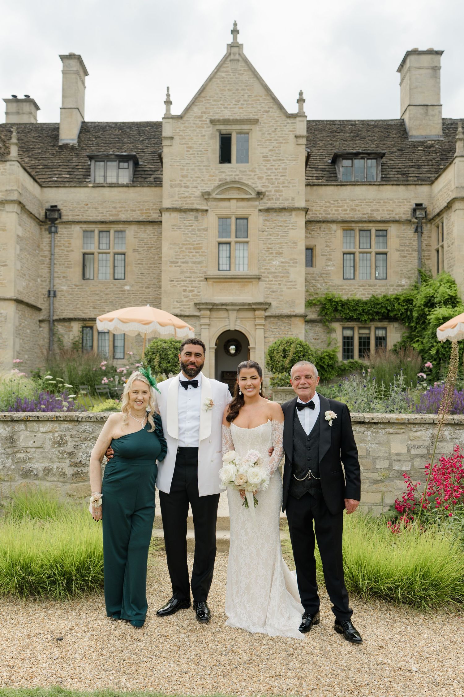 Wedding couple poses with family in front of grand stone manor house at English countryside wedding.
