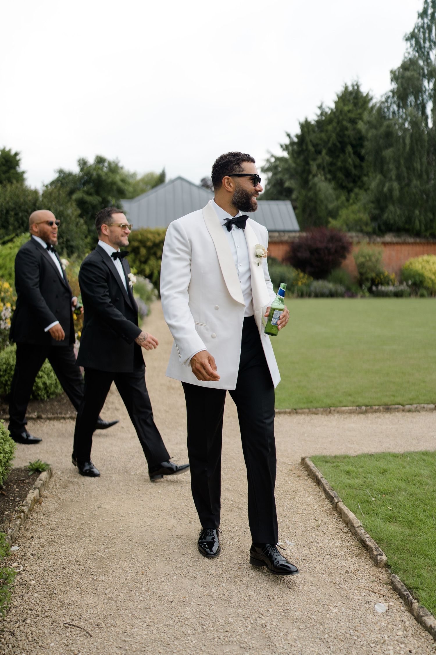 Groom in white tuxedo jacket walks garden path with groomsmen in black tuxedos at outdoor wedding.