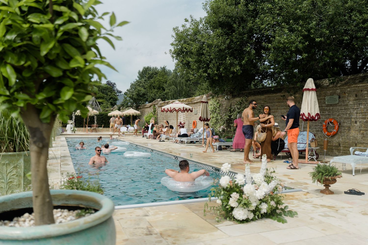 Wedding guests relax and swim in outdoor pool at elegant stone manor venue on sunny day.