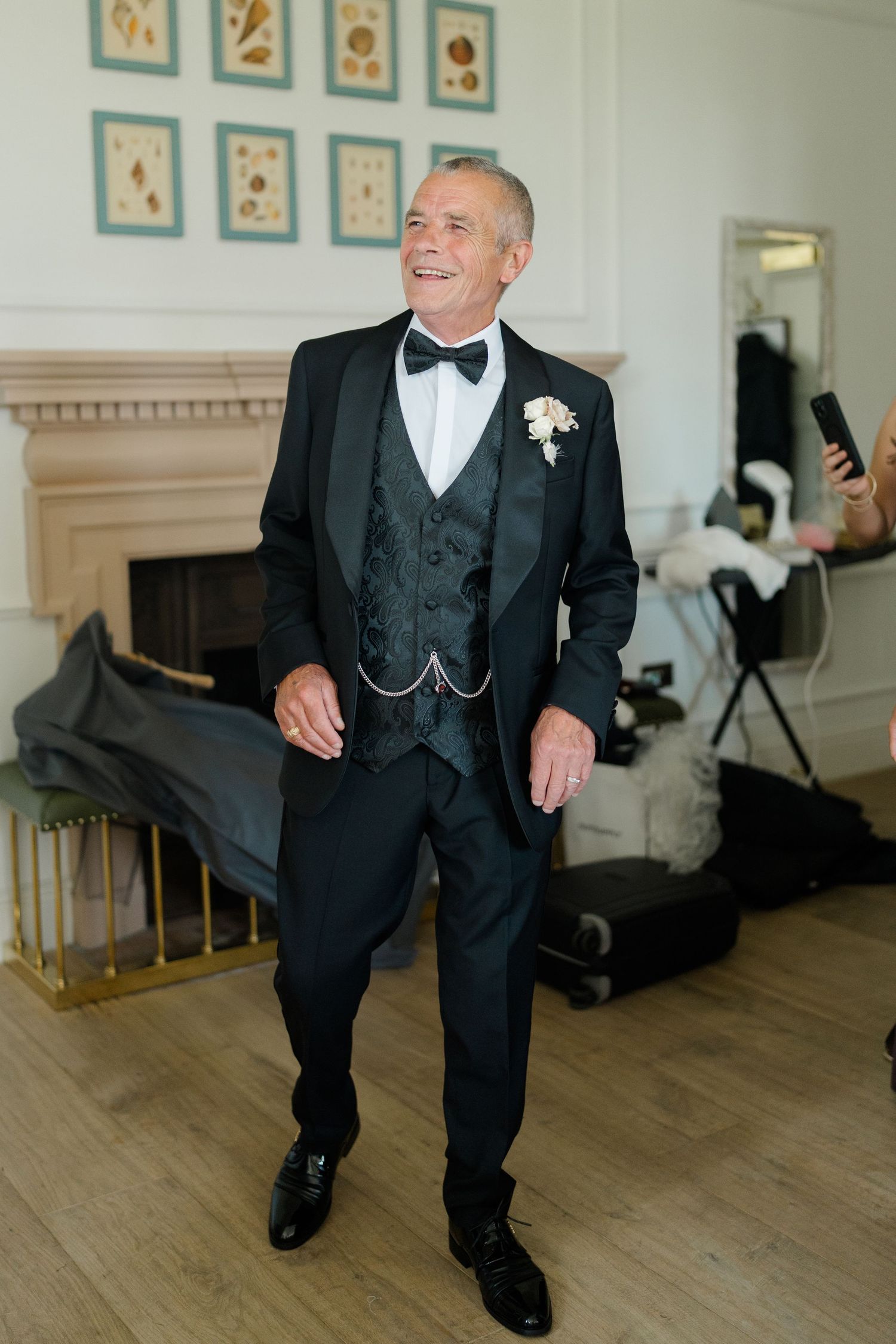 Smiling groom in black tuxedo with bow tie and boutonniere standing by fireplace on wedding day.