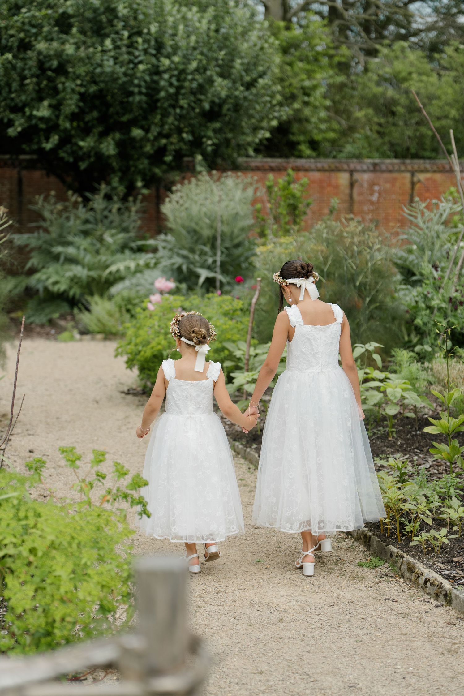 Two flower girls in white dresses walk hand in hand through lush garden.