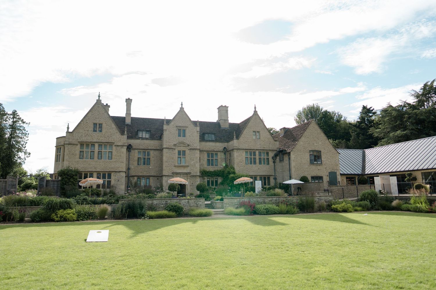 Grand Cotswold stone manor house with manicured lawn and lush gardens on a sunny day.