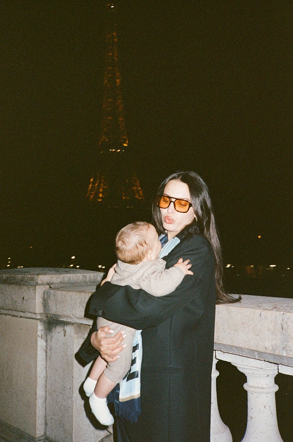 Woman holding a baby in front of the Eiffel Tower at night in Paris, wearing orange sunglasses and a black coat.