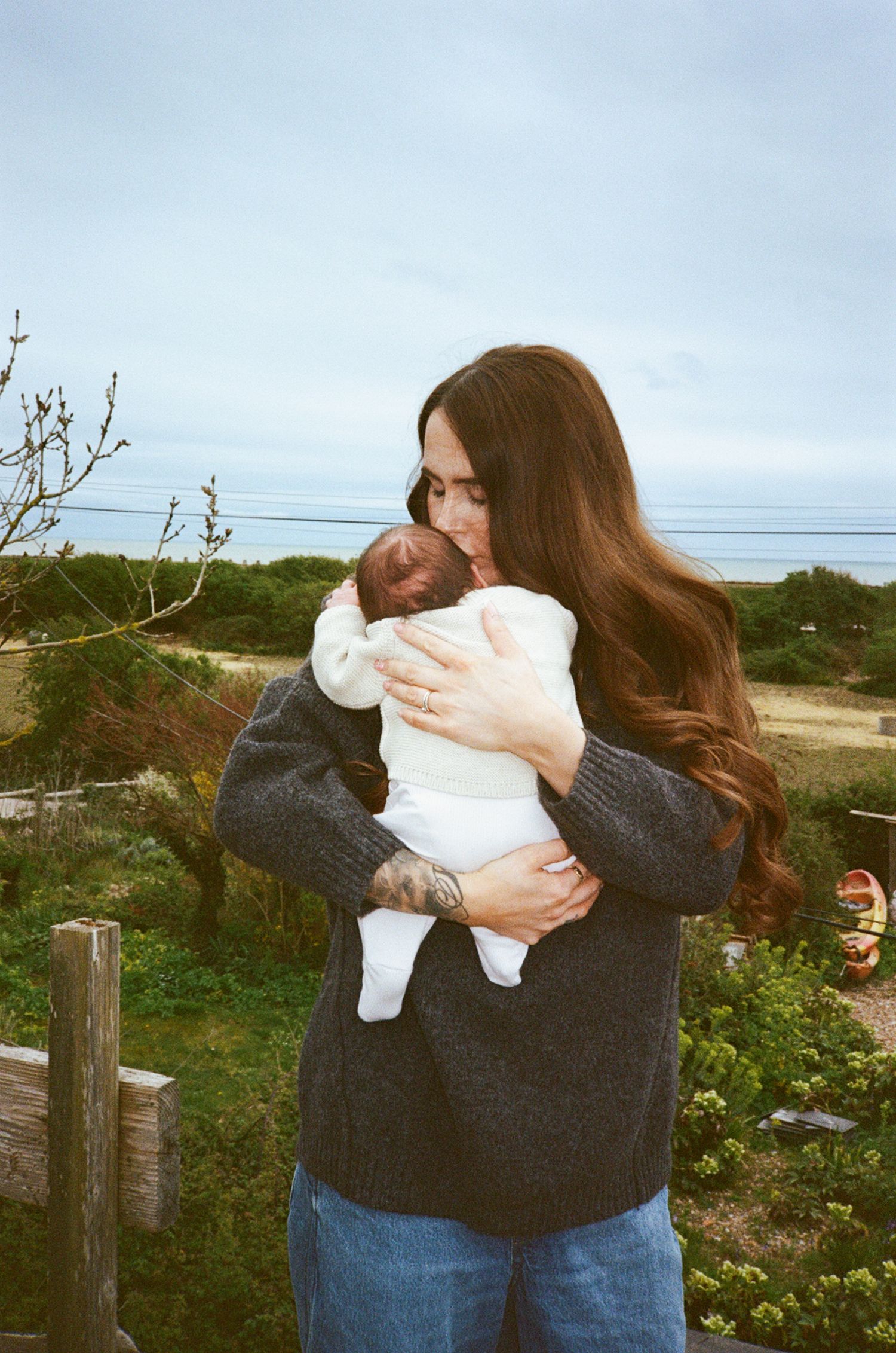 A woman with long brown hair holds a newborn baby outdoors, wearing a gray sweater with a tattoo visible.