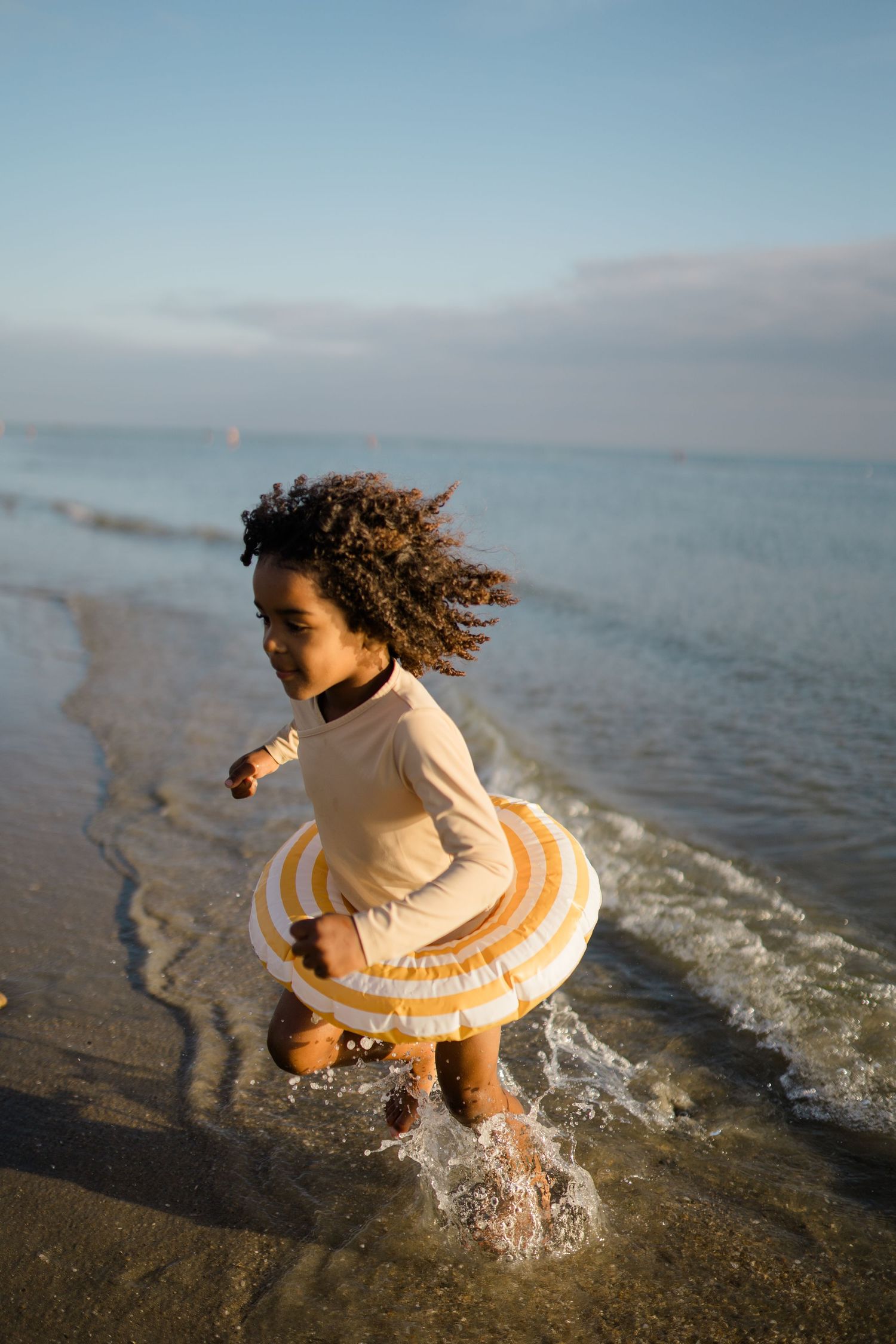 A curly-haired figure in a white dress splashes joyfully in the ocean waves at sunset.