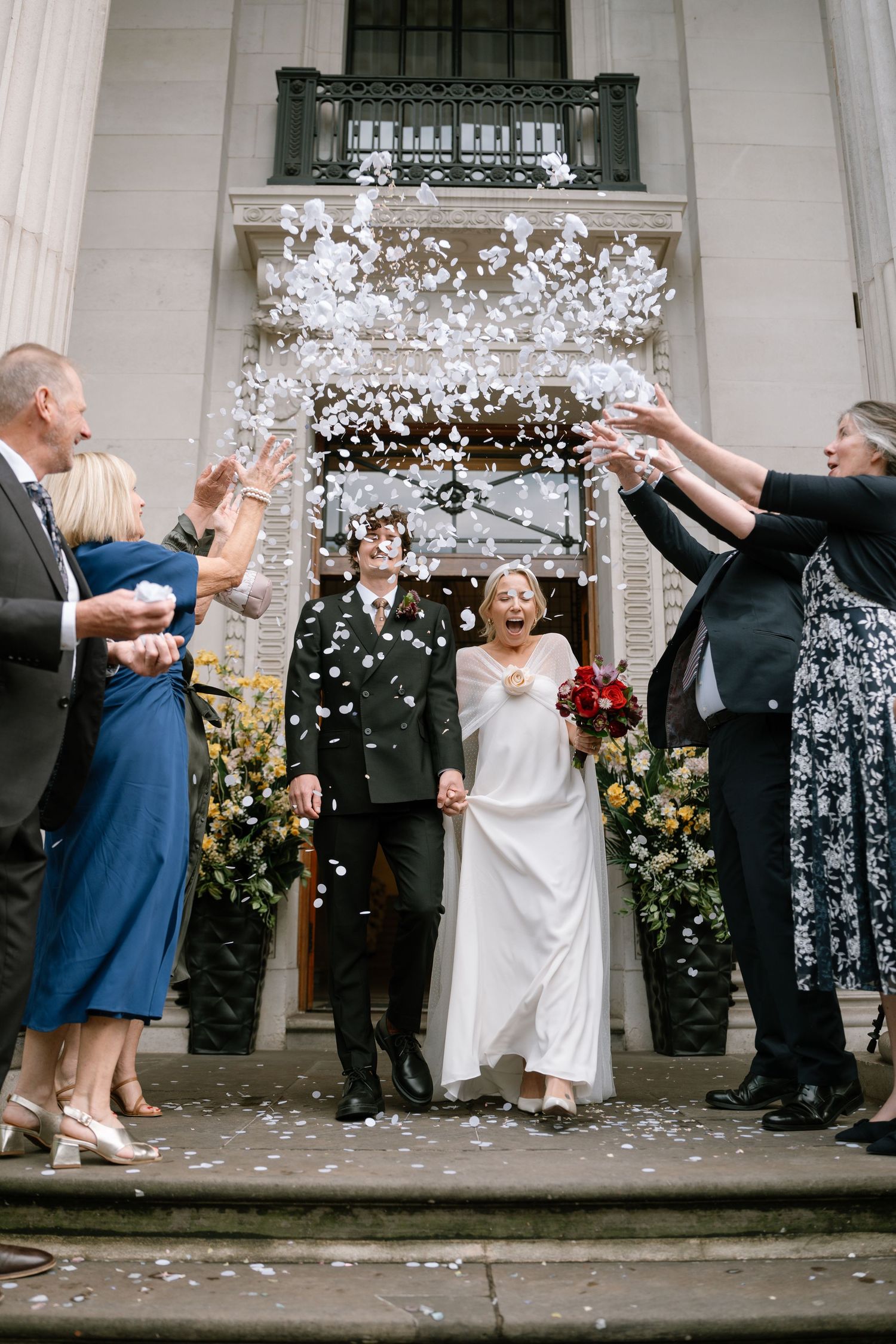Wedding party throws confetti in joyful celebration on the steps of a classical building.