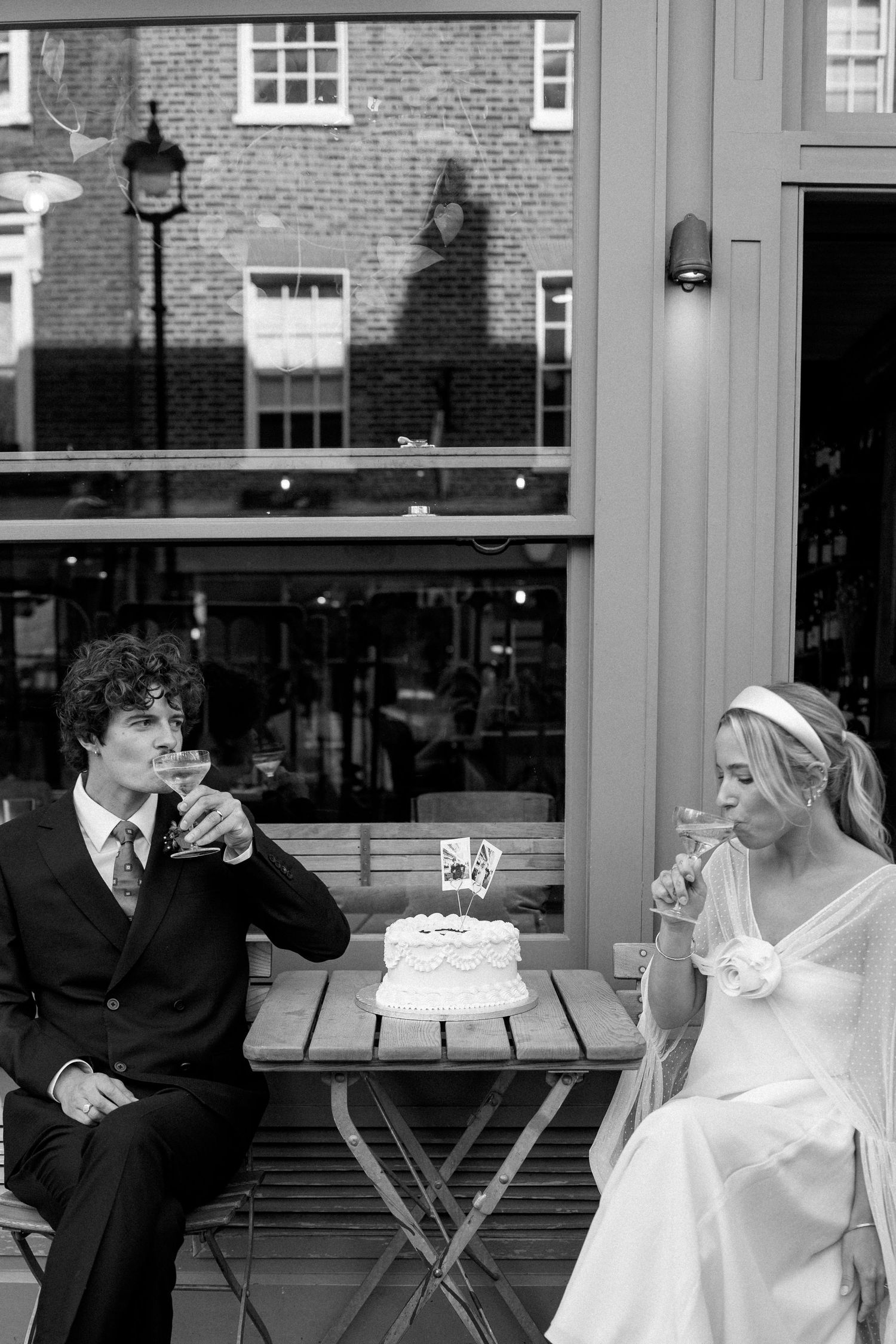 A couple enjoys beverages beside their wedding cake at an outdoor cafe setting.
