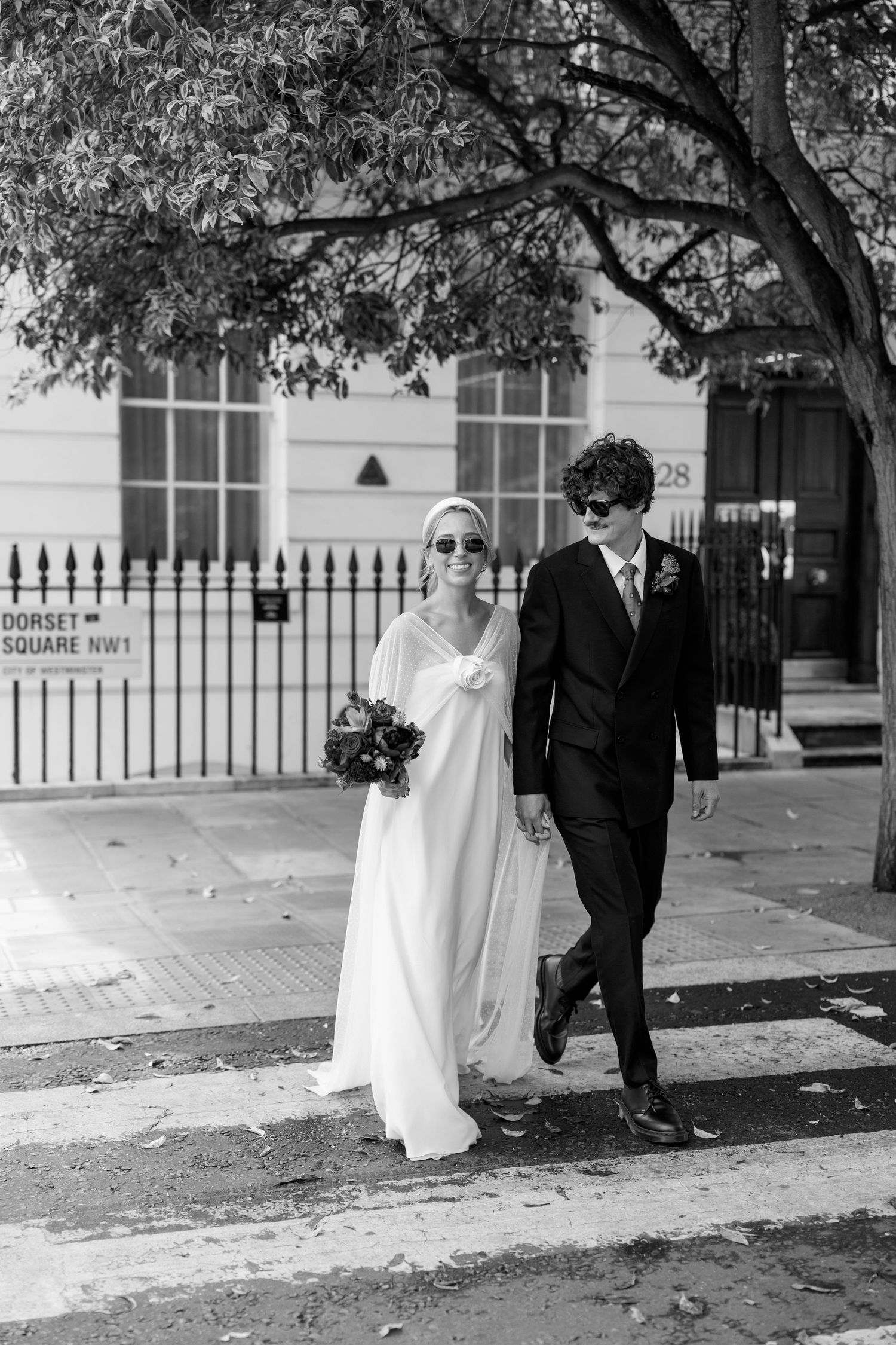 A couple strolls along a sidewalk with iron fencing and trees in an elegant London neighborhood.