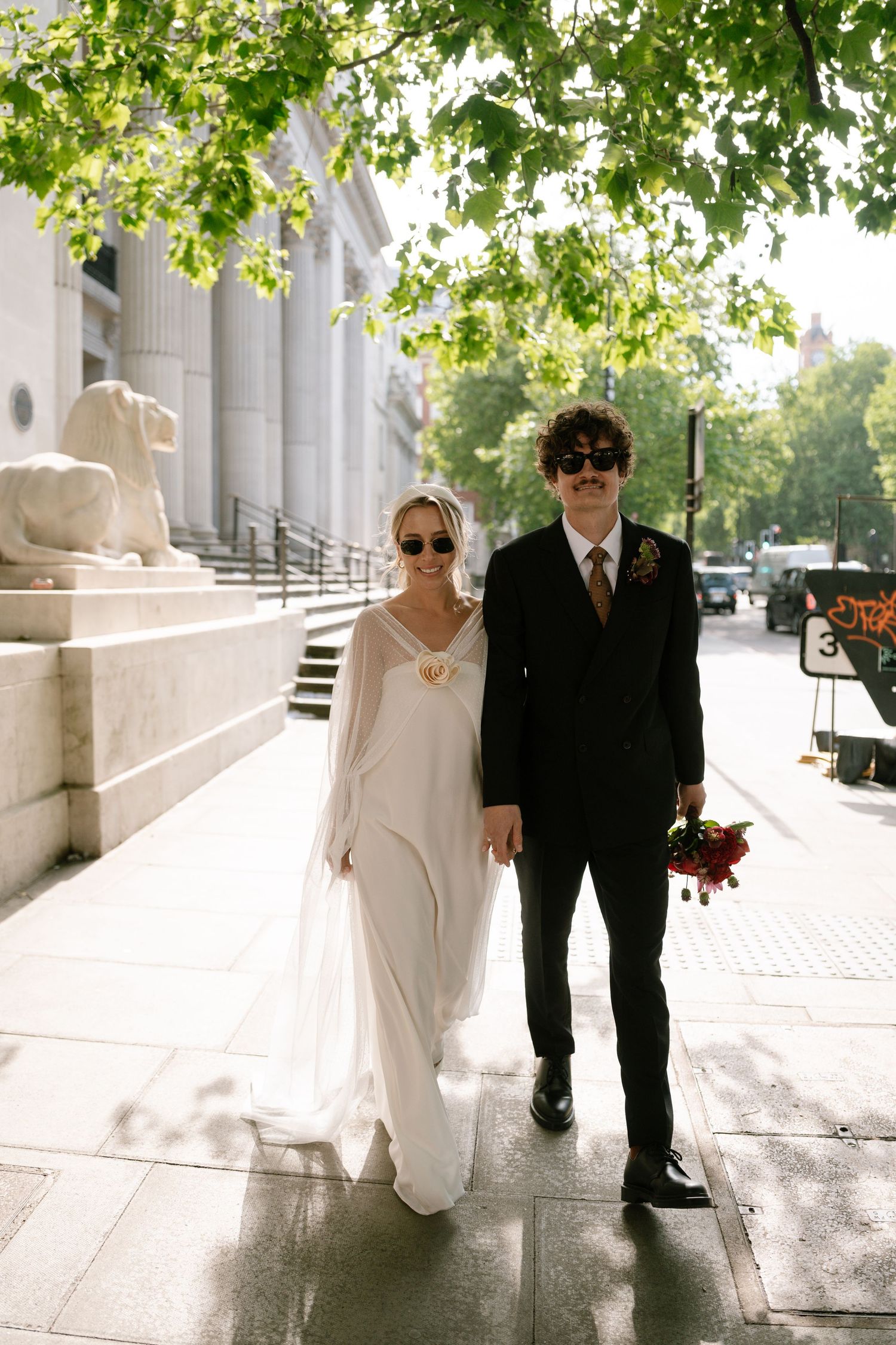 A couple walks along a sunlit path beside a classical building with columns.