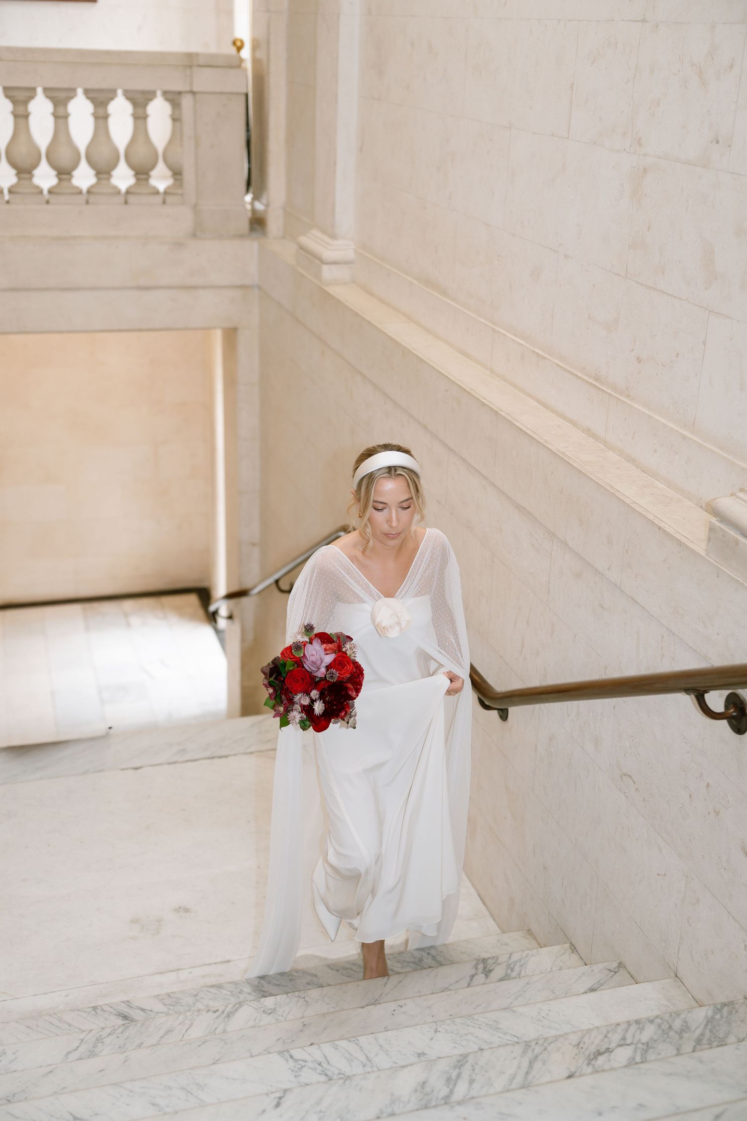Bride descending marble stairs holding a red bouquet in an elegant white interior setting.