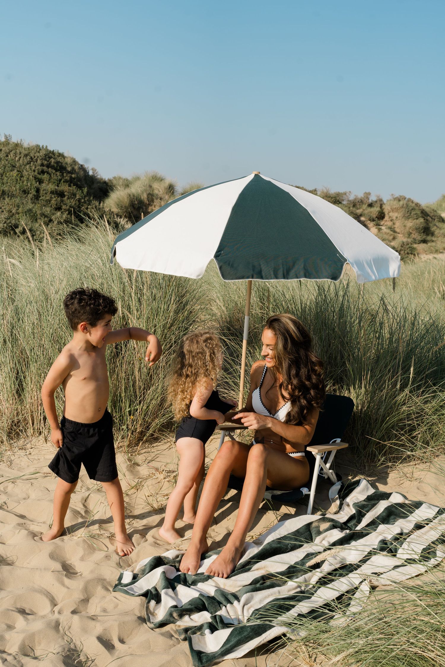 Group relaxing on striped beach blanket under white umbrella among coastal dunes.