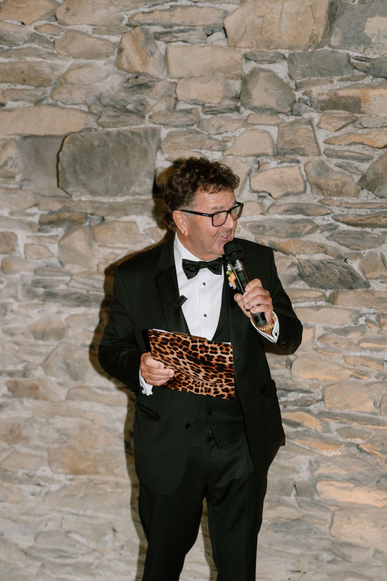 Person in formal wear with glasses and bowtie speaking at an event with stone wall backdrop.