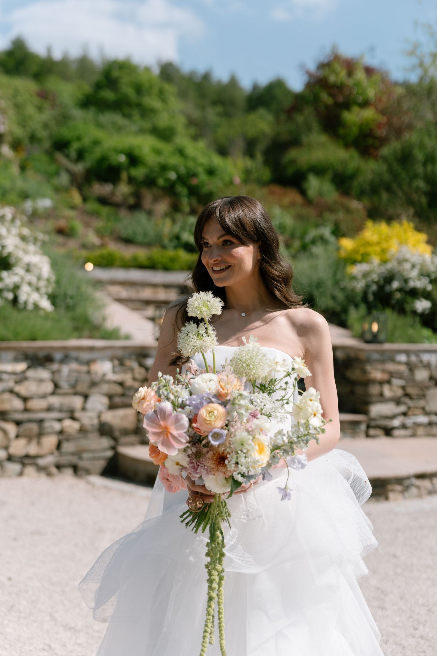 A bride in a strapless gown poses with her bouquet in a garden with stone walls.