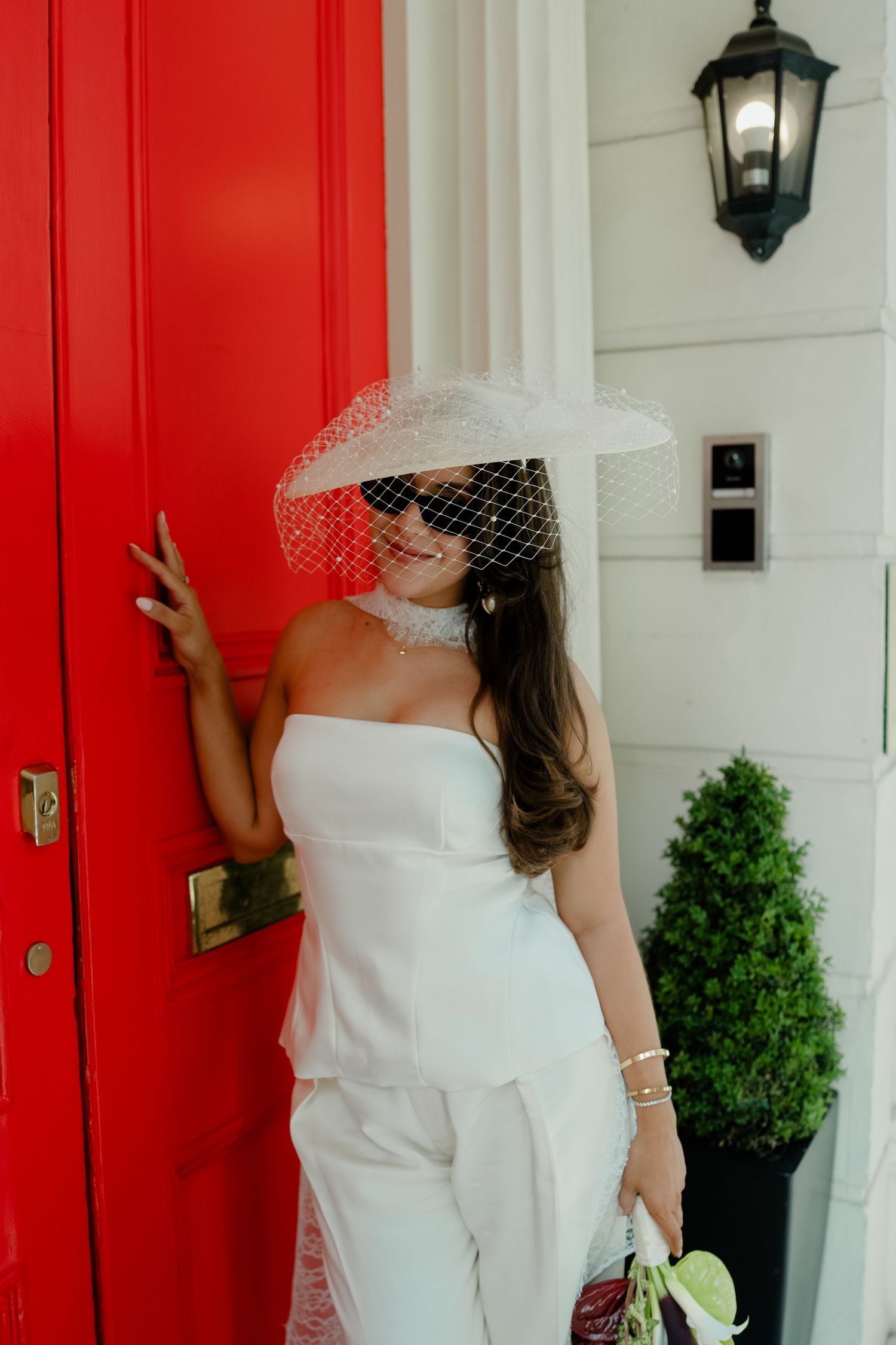 Stylish bride in white strapless jumpsuit and wide-brim veil hat posing by a bold red door with sunglasses.