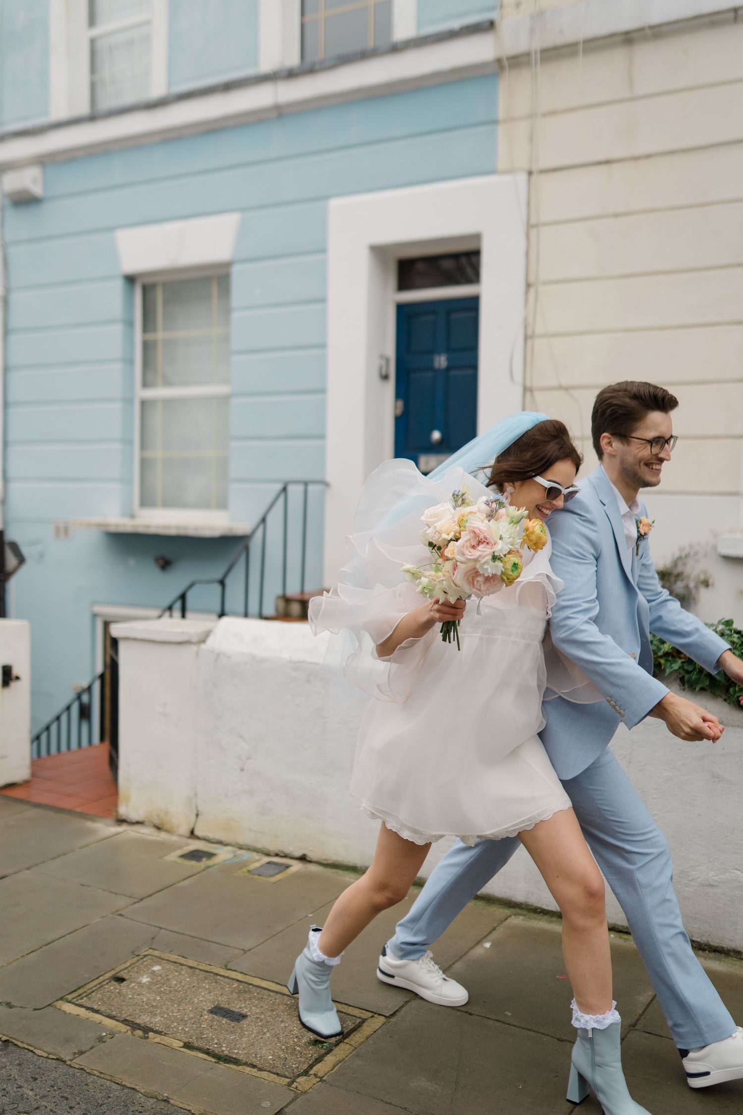 Bride in short white dress and groom in light blue suit walking joyfully on a colorful street in Notting Hill, London.