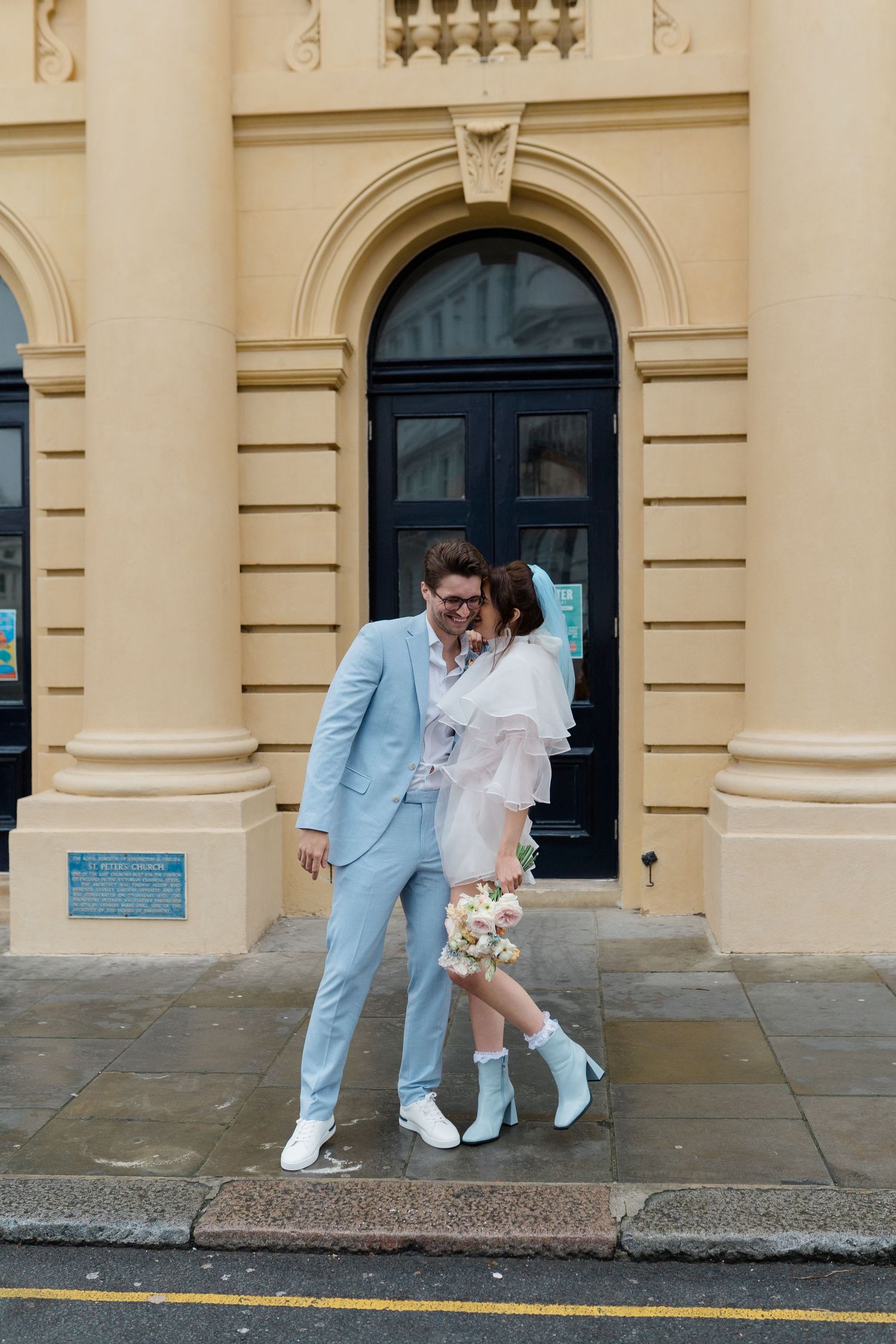 Couple in matching white boots pose outside a yellow building, bride in short white dress holding bouquet, groom in light blue suit.