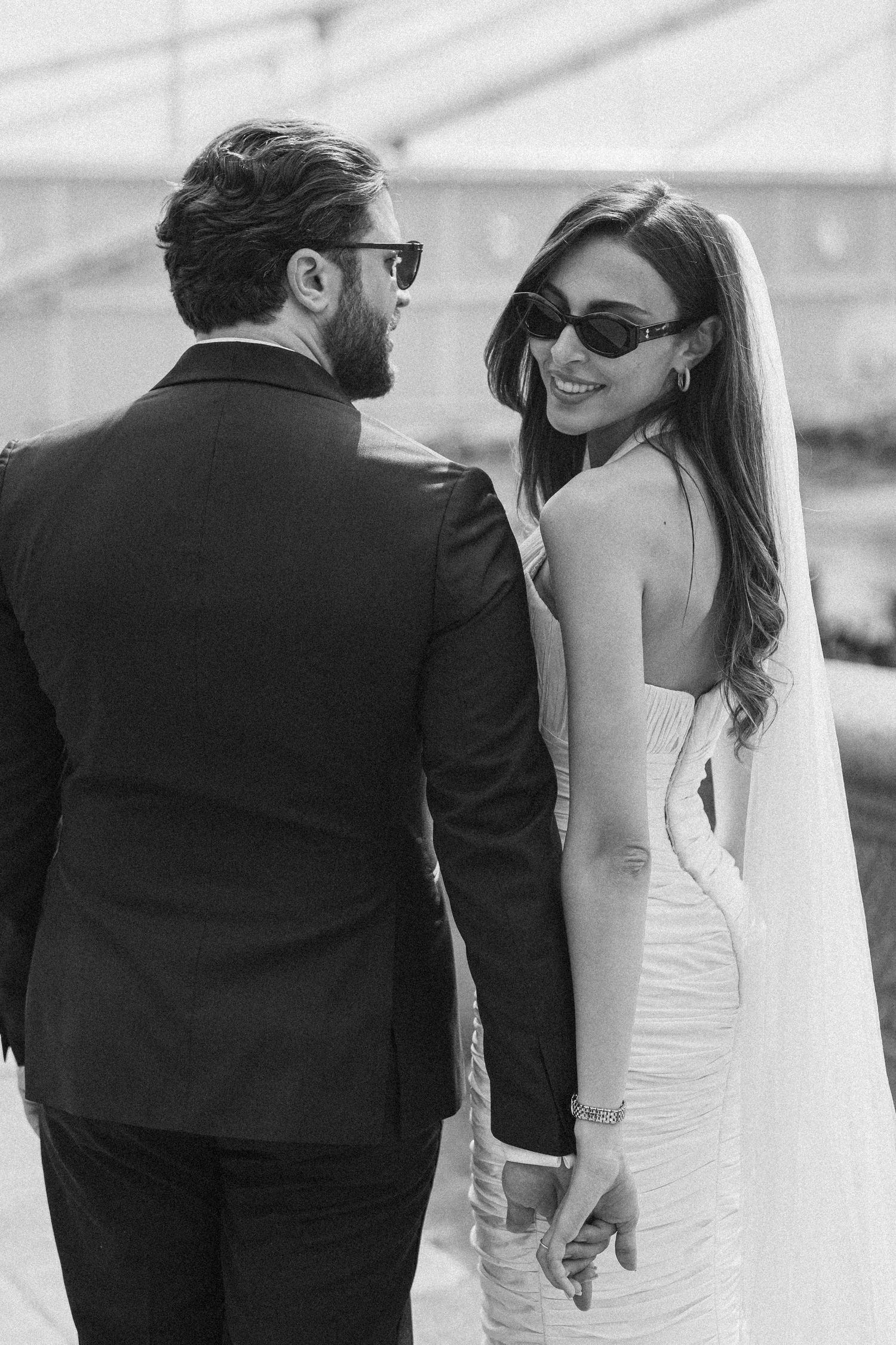 Black and white photo of a smiling bride in backless gown and veil holding hands with groom in suit outdoors.