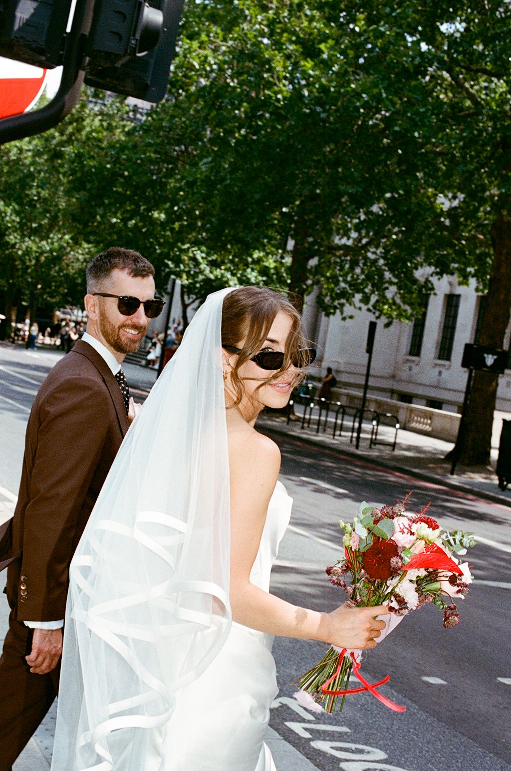 Bride in white veil and sunglasses holding red bouquet with groom in brown suit on street.