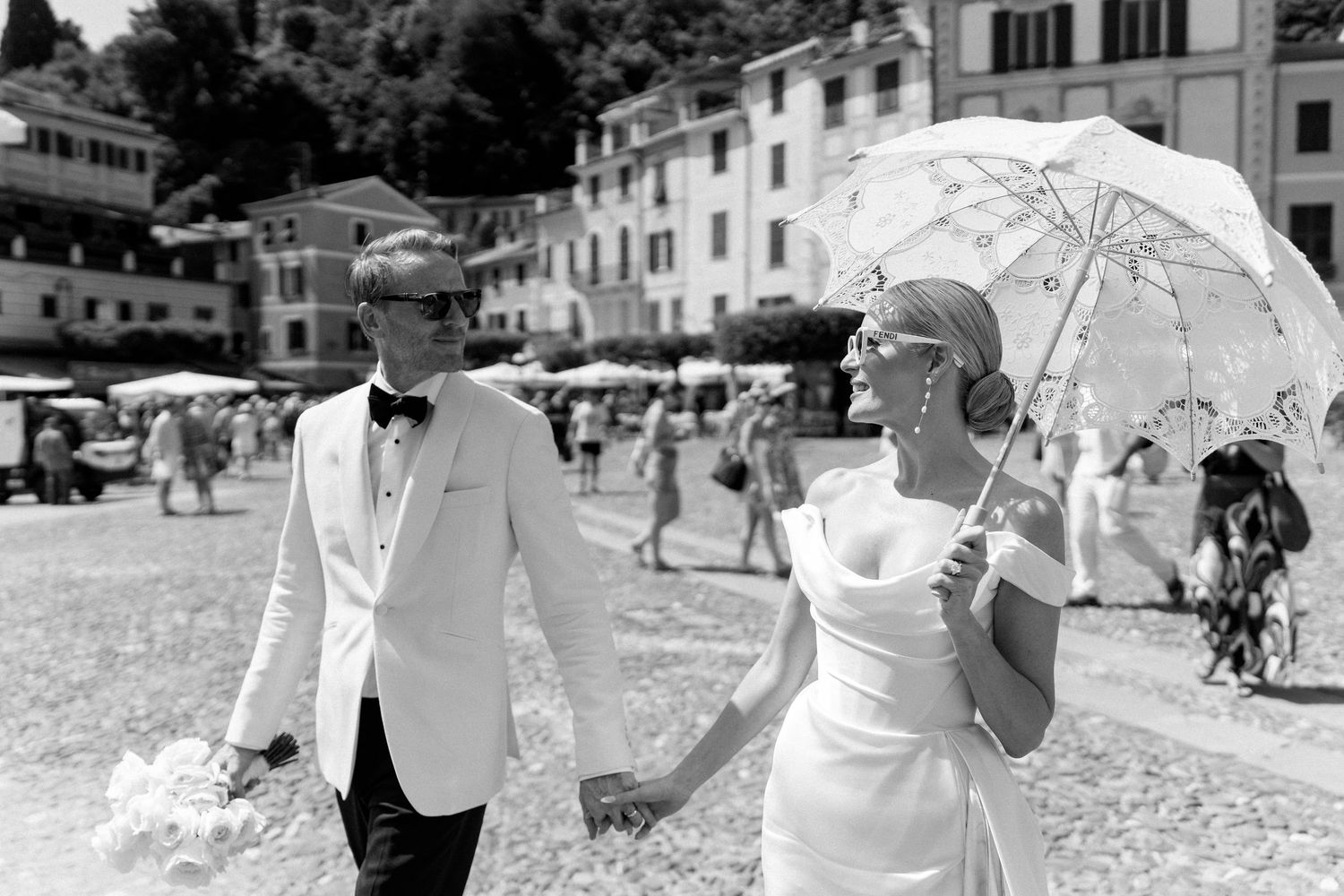Bride and groom walking hand in hand through Portofino square, bride holding lace parasol.