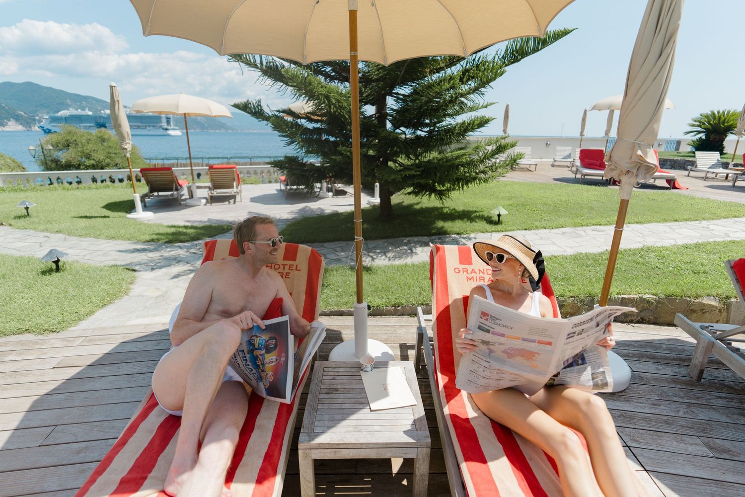 Two guests relaxing on striped sun loungers reading magazines at seaside resort under umbrella.