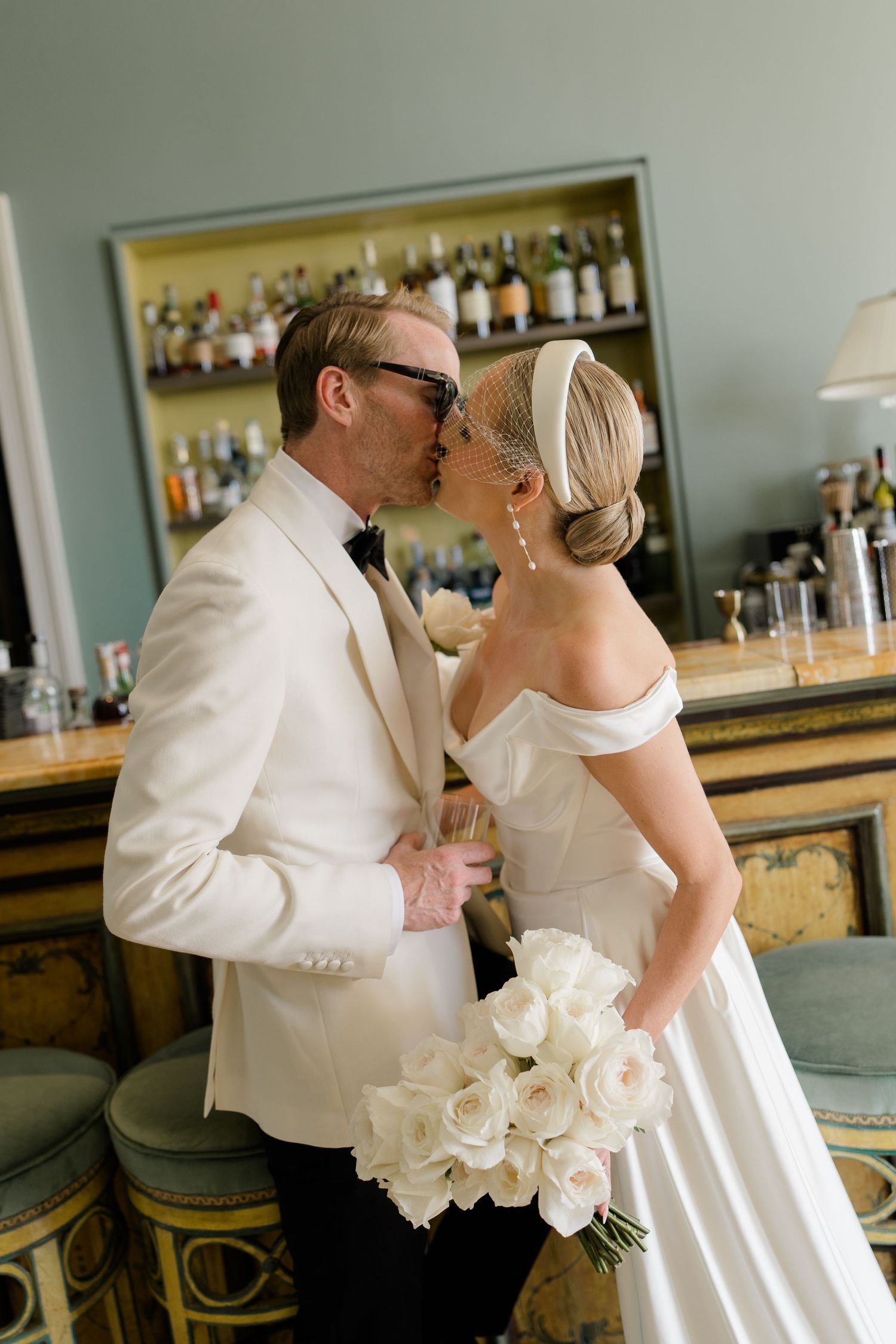 Bride and groom share a kiss at bar, she holds white rose bouquet, he wears white tuxedo jacket.