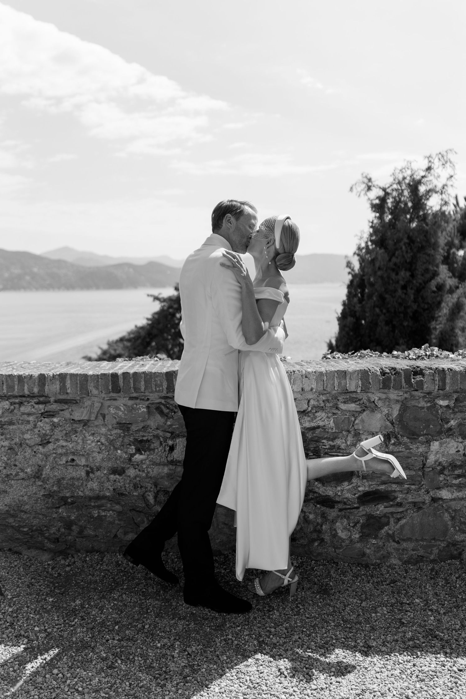 Couple sharing a romantic kiss by a stone wall overlooking the sea and mountains in a black and white photo.