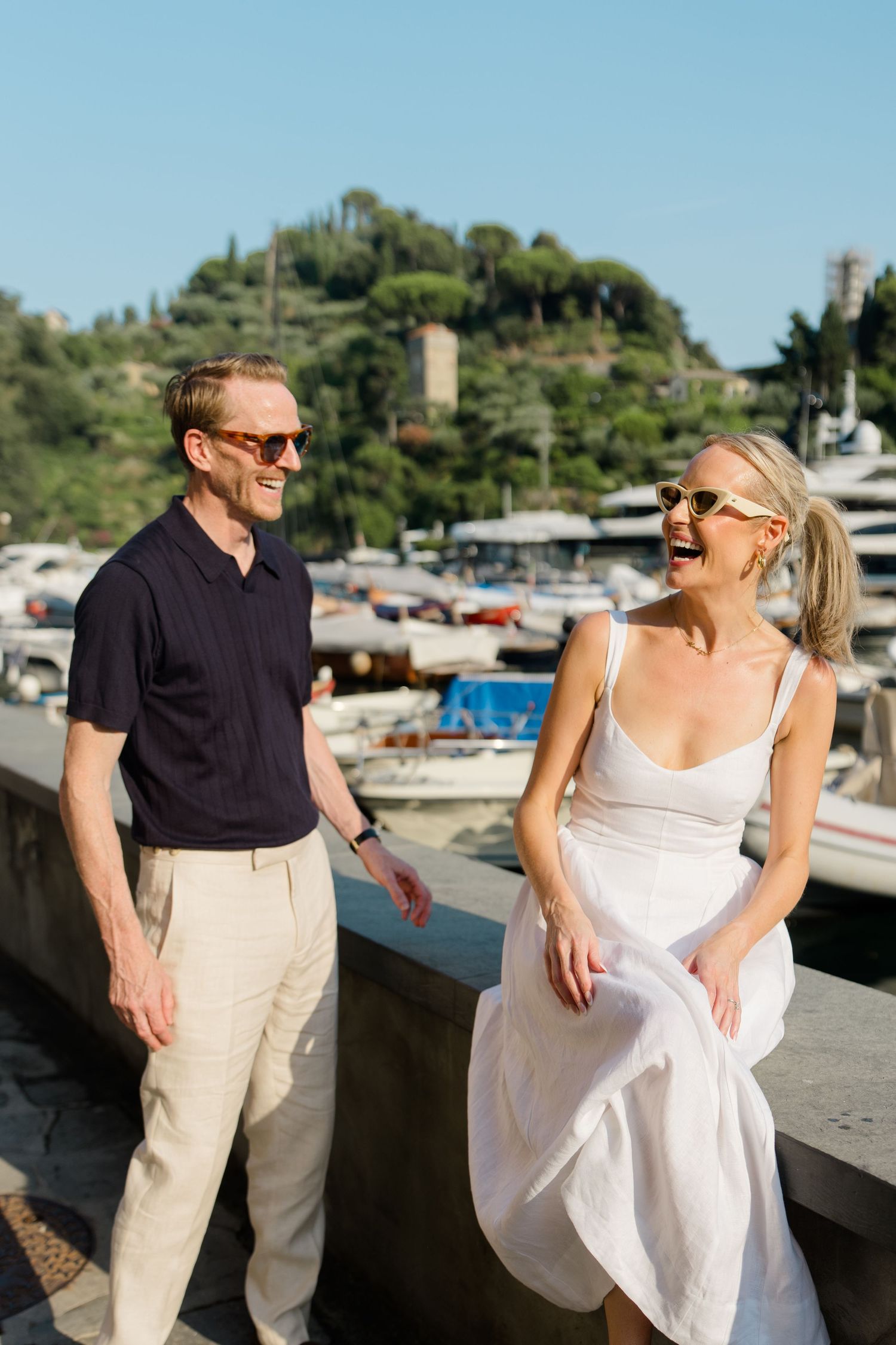 Couple laughing together by a marina in Portofino, Italy, wearing sunglasses on a sunny day.